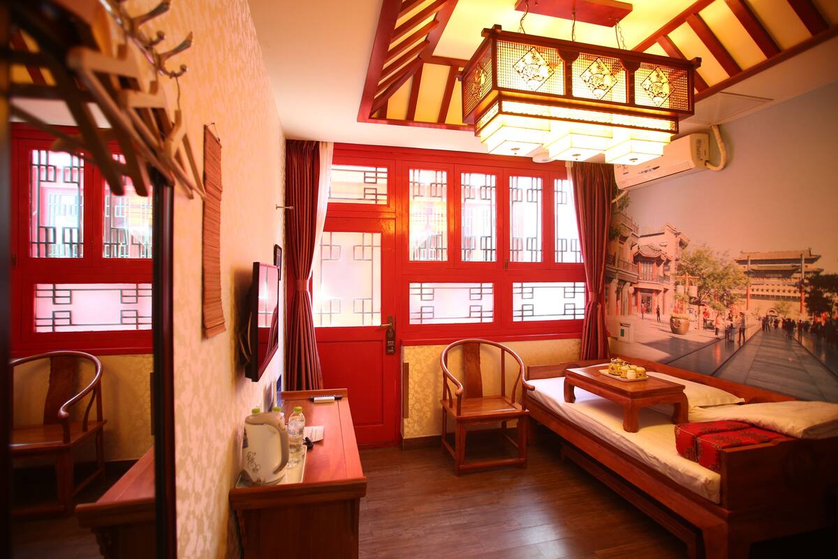A traditional room with a wooden sofa, a small table, and a chair. There are two windows with red frames and a mural on the wall depicting a street scene.