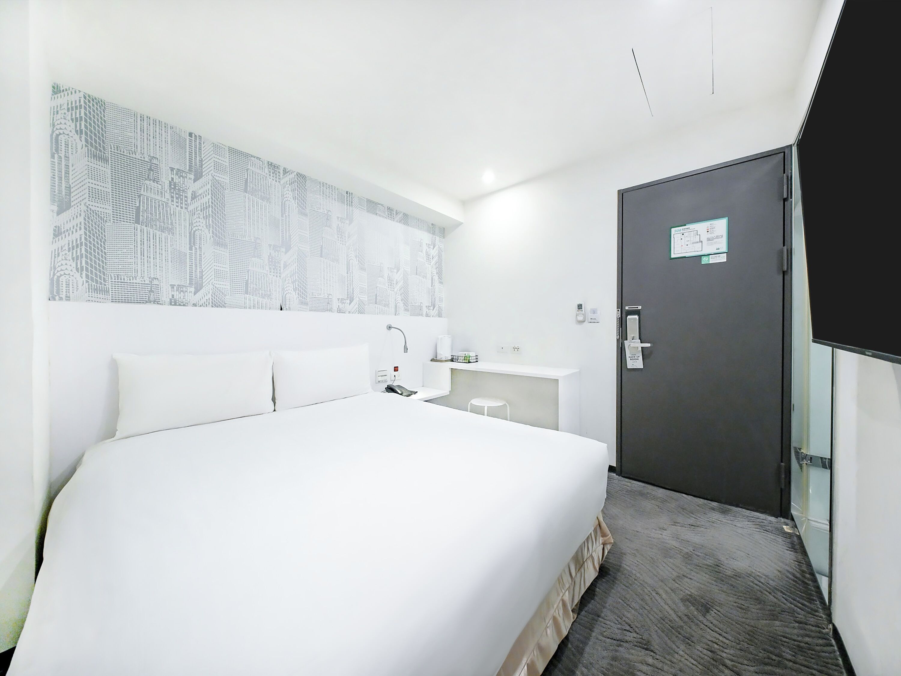 A modern hotel room with a large bed, a small bedside table, a mirror, a wall-mounted sink, and a door with a lock.
