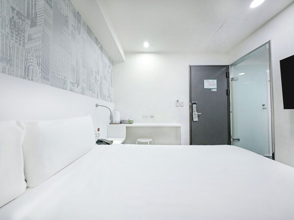 Habitación de hotel con cama, mesita de noche, espejo, teléfono de pared y puerta con panel de vidrio.