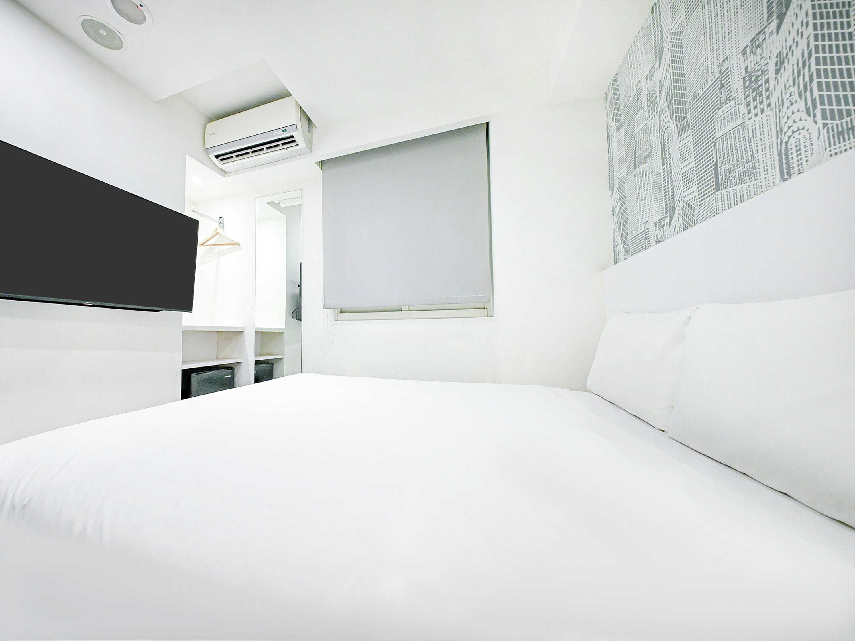A modern hotel room with a large bed, a flat-screen TV, a window with a view of buildings, and an air conditioning unit.