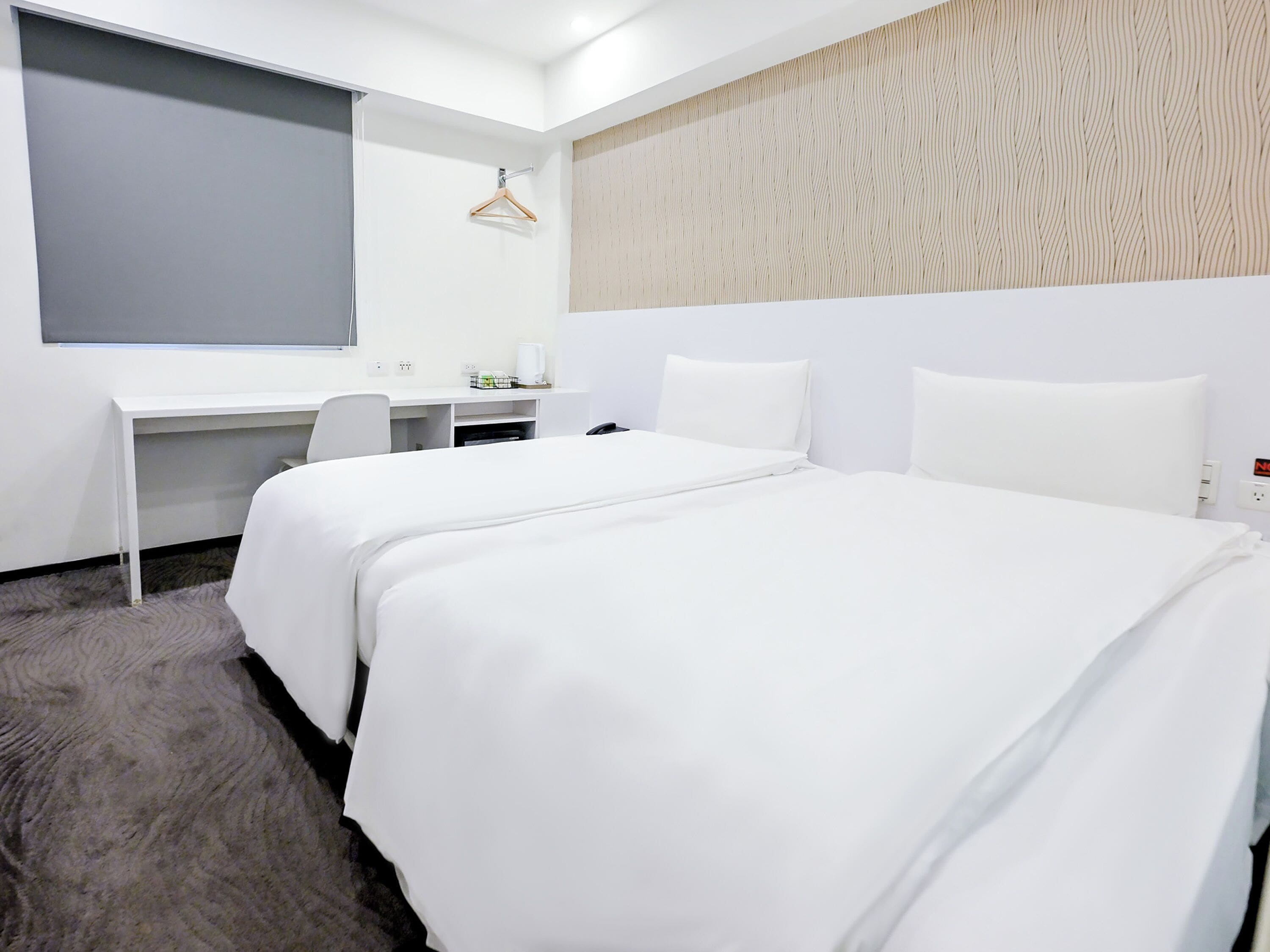 Ein Hotelzimmer mit Bett, Schreibtisch, Stuhl, Kleiderhaken und einem großen Fenster.
