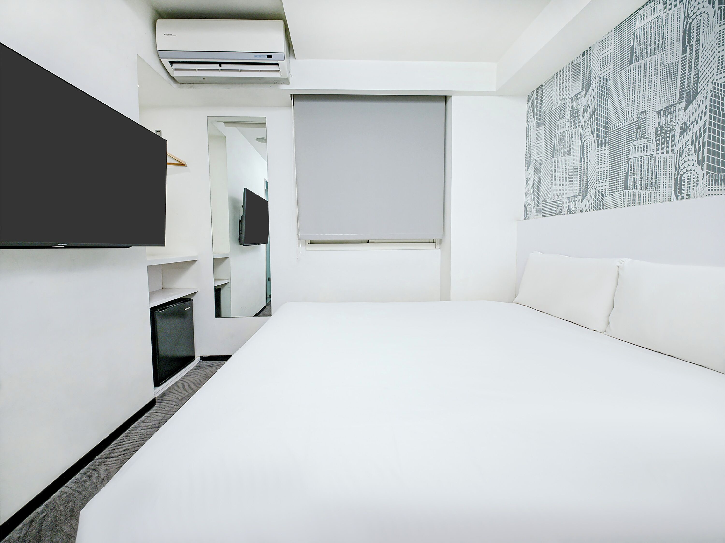 A modern hotel room with a bed, a flat-screen TV, an air conditioning unit, a window with blinds, and a patterned wall.