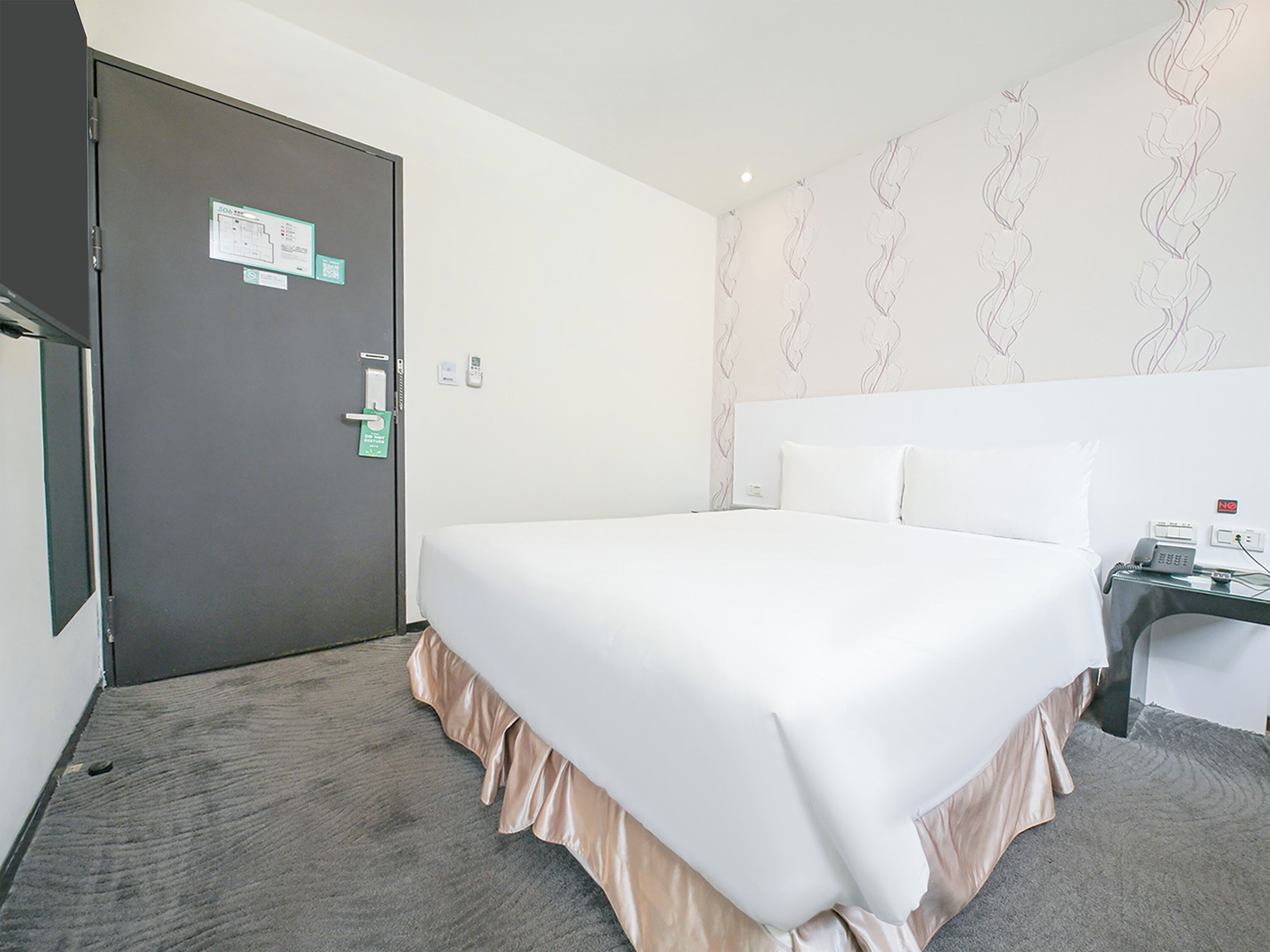 Une chambre d’hôtel avec un lit, une table de chevet avec un téléphone, une porte dotée d’un lecteur de carte magnétique et un mur orné d’un motif floral.