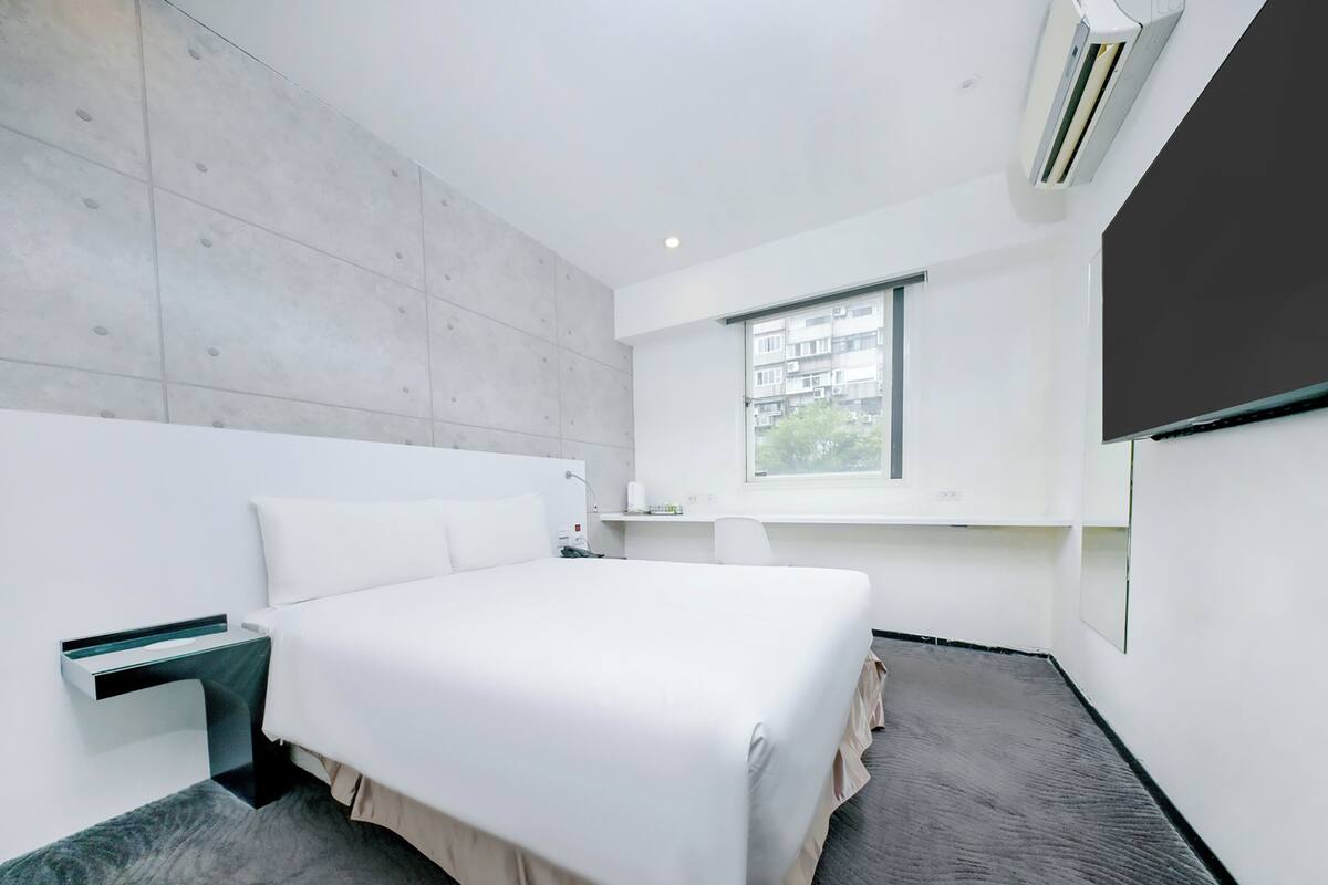 A modern hotel room with a large bed, a bedside table, a small desk, a window with a view, and an air conditioning unit.