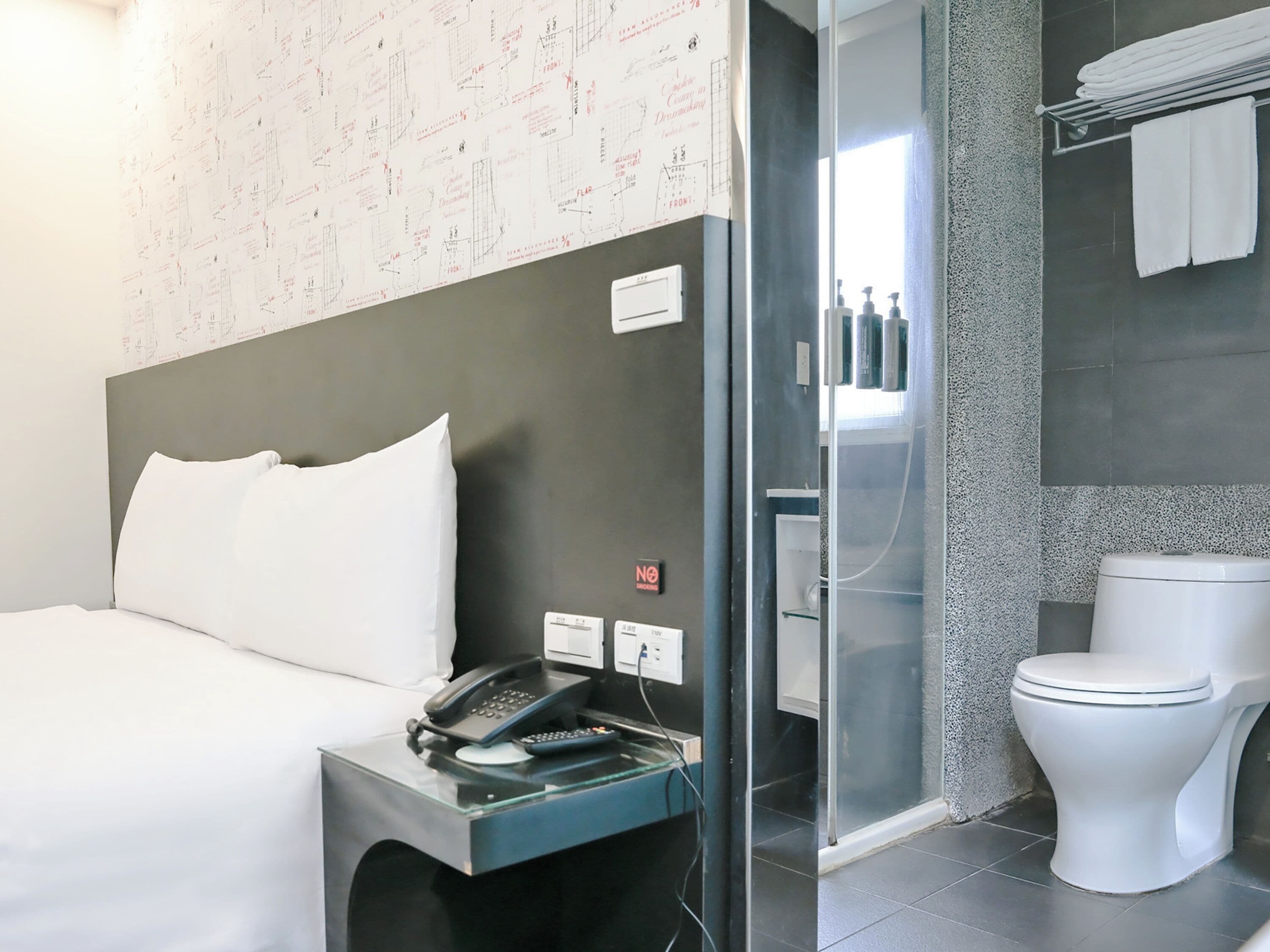 A hotel room with a bed, a bedside phone, a glass shower enclosure, a toilet, and a towel rack.