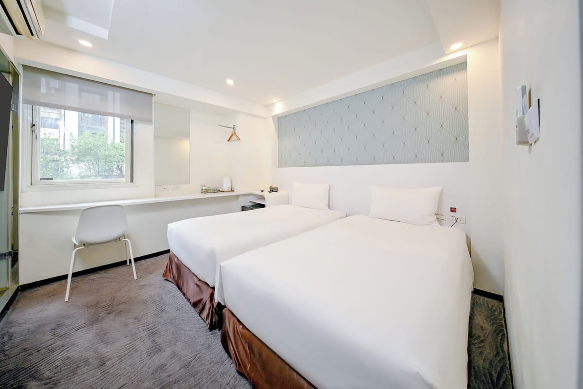 A hotel room with two beds, a desk, a chair, a window, and a wall-mounted light fixture.