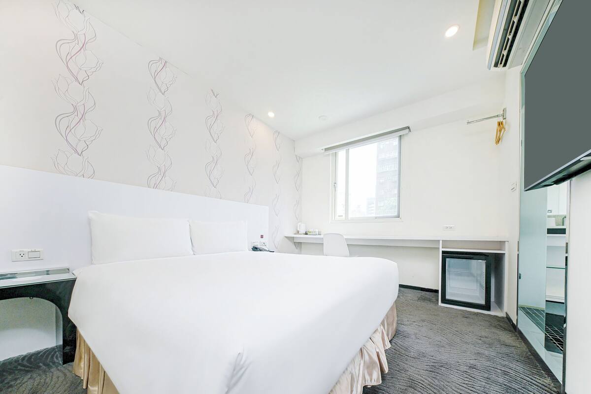 A hotel room with a large bed, a desk, a chair, a television, and a window.