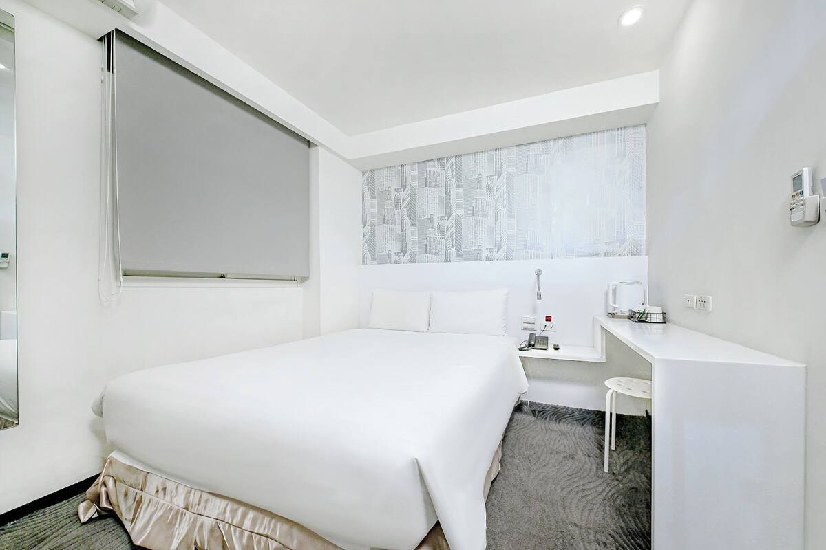 A hotel room with a large bed, a desk, a chair, and a window with a cityscape view.