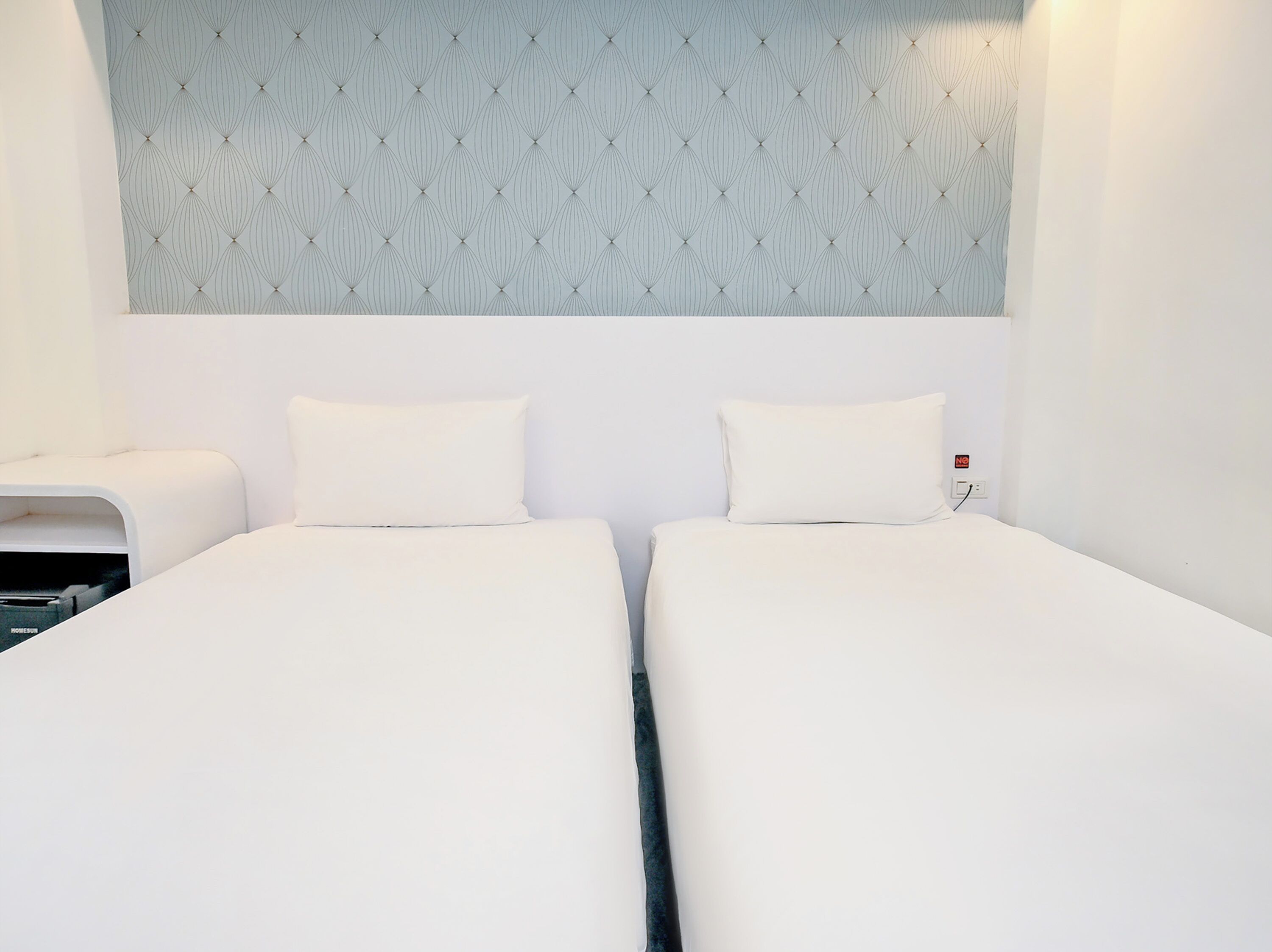 Habitación doble con dos camas, cabecero con estampado y enchufe de pared.