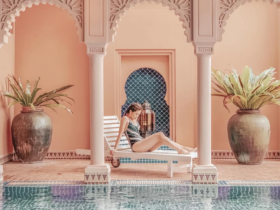 A pool area with a person relaxing on a lounge chair, surrounded by ornate arches and large potted plants.