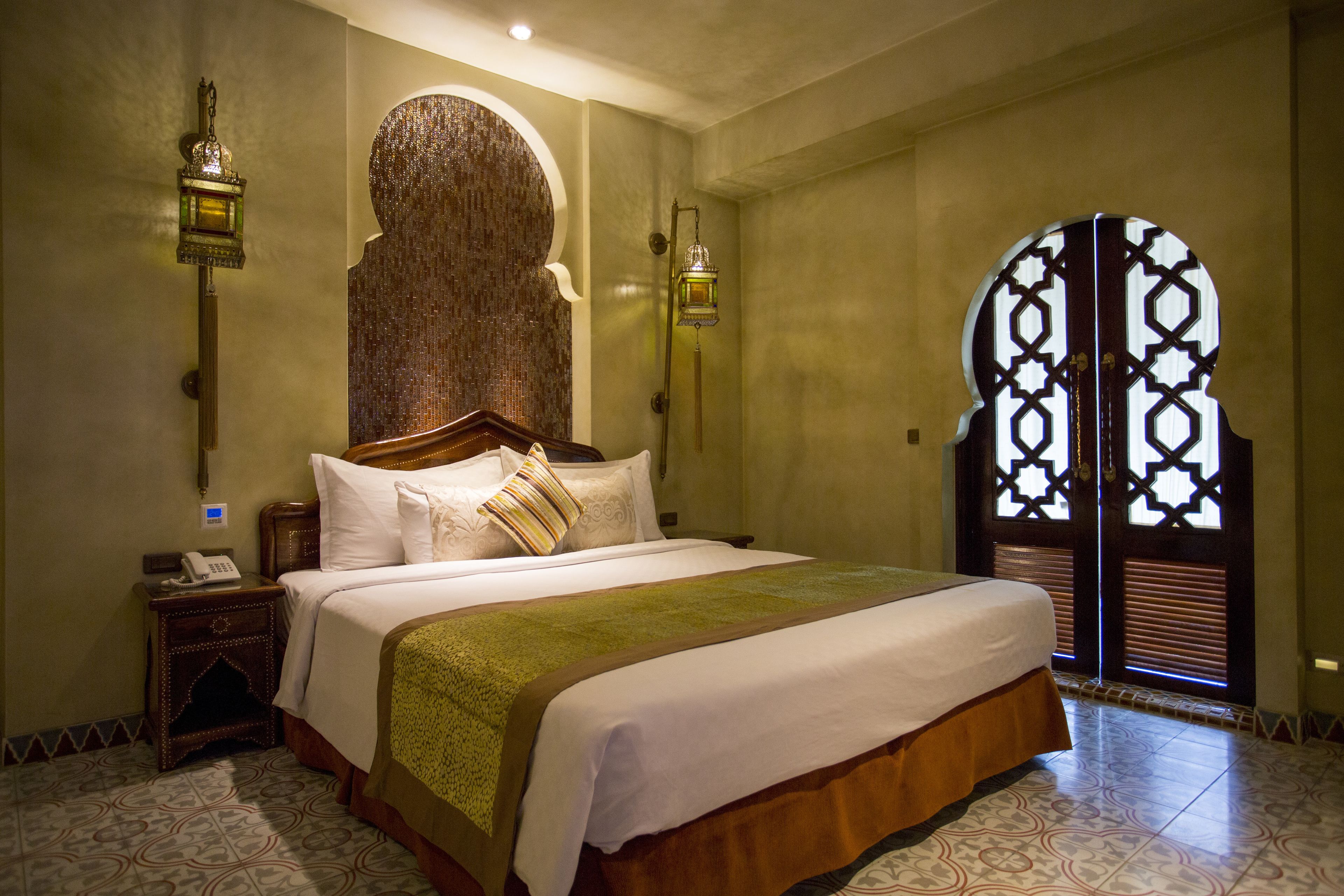 A well-appointed hotel room with a large bed, a nightstand with a telephone, decorative wall sconces, and a unique arched doorway.