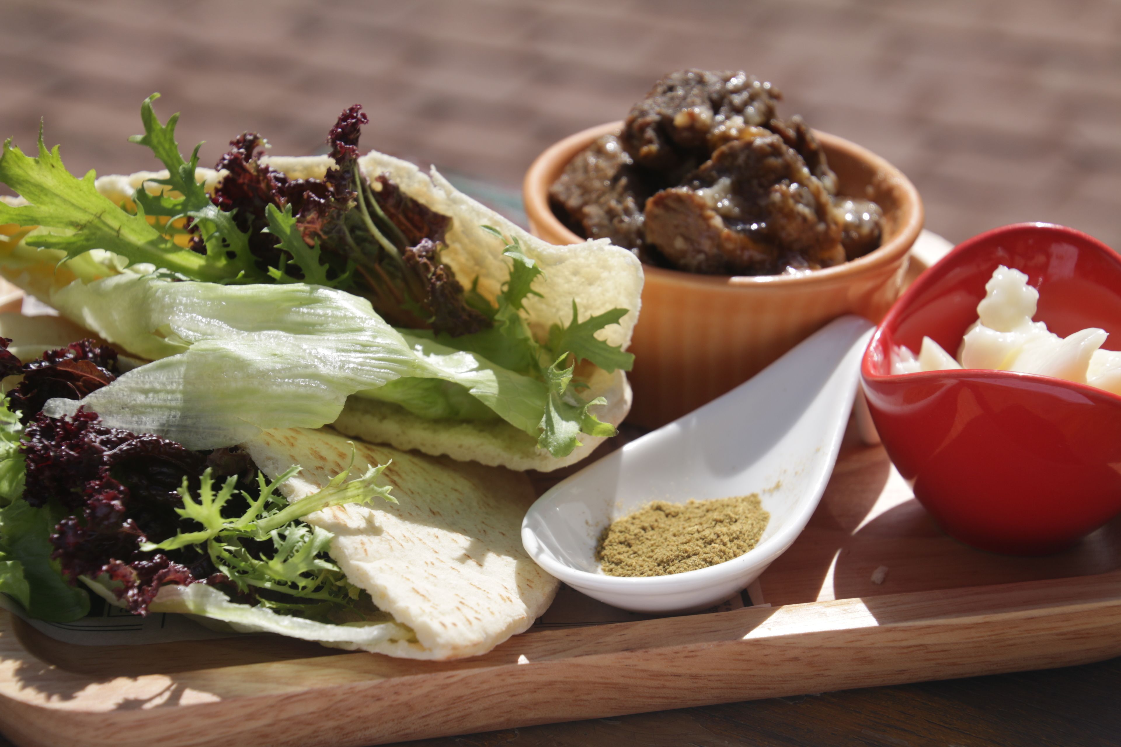 A wooden tray with a wrap containing lettuce, a bowl of meat, a small bowl of seasoning, and a red bowl with a white substance.
