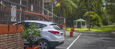Un estacionamiento con un coche blanco estacionado junto a un edificio de fachada de ladrillo y techo verde. Se ven plantas en macetas y un pabellón al fondo.