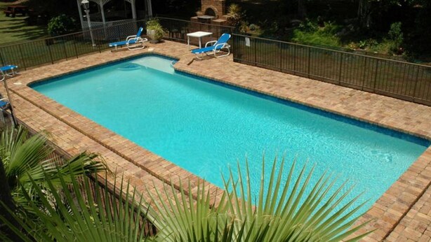 Una piscina rectangular con borde de ladrillo, rodeada por una cerca de metal.