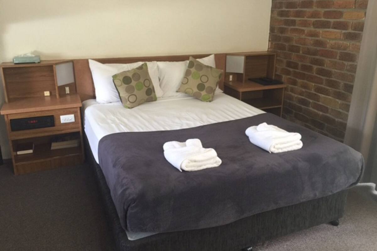 A neatly made bed with a dark bedspread and two folded towels on it. There are two pillows with a polka dot pattern and a wooden nightstand with a small lamp.