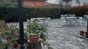 Een terras buiten met stenen vloer, witte metalen tafels en stoelen, en potplanten.