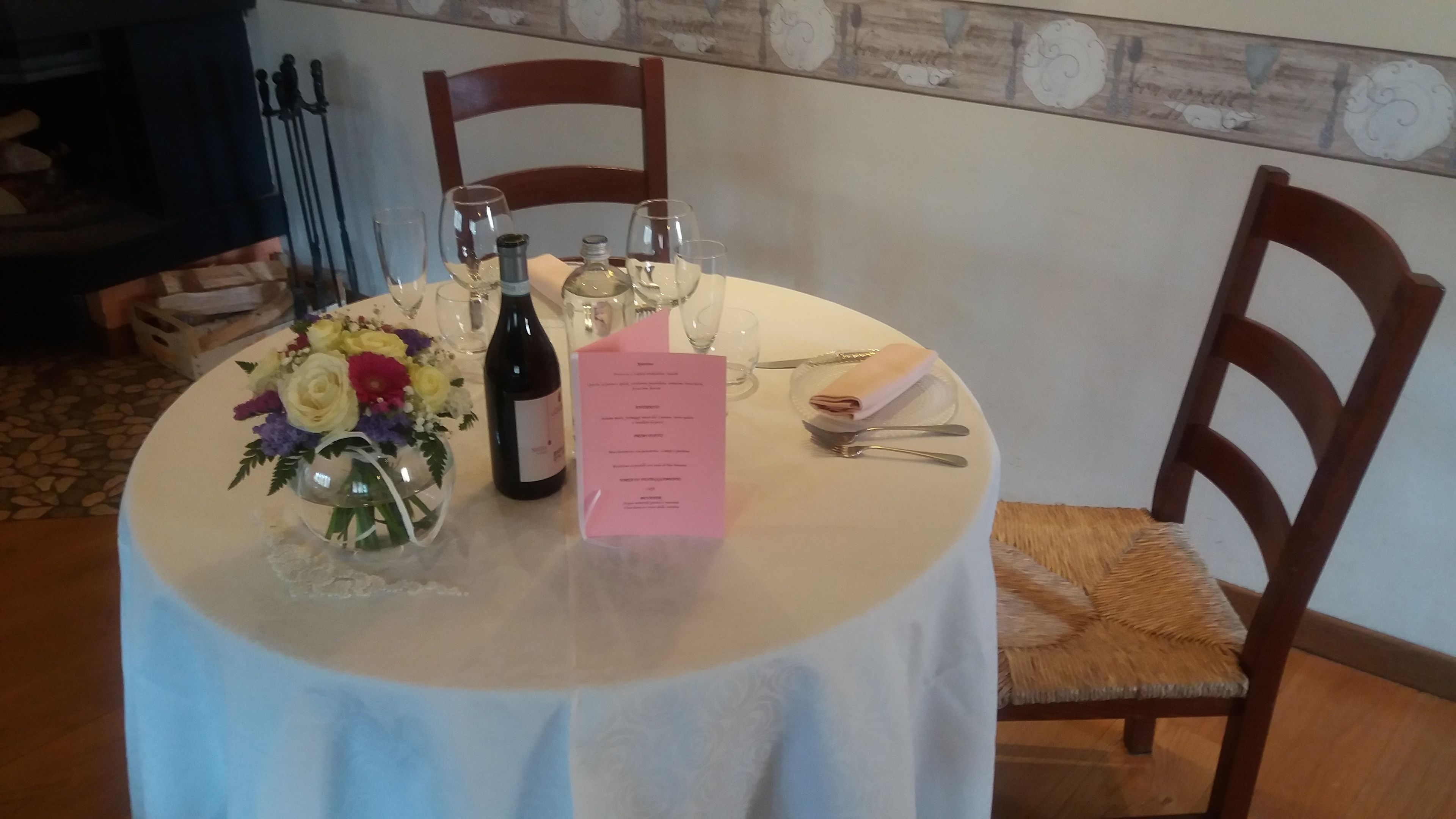 A round dining table with a white tablecloth, a menu, wine glasses, a bottle of wine, and a floral centerpiece.