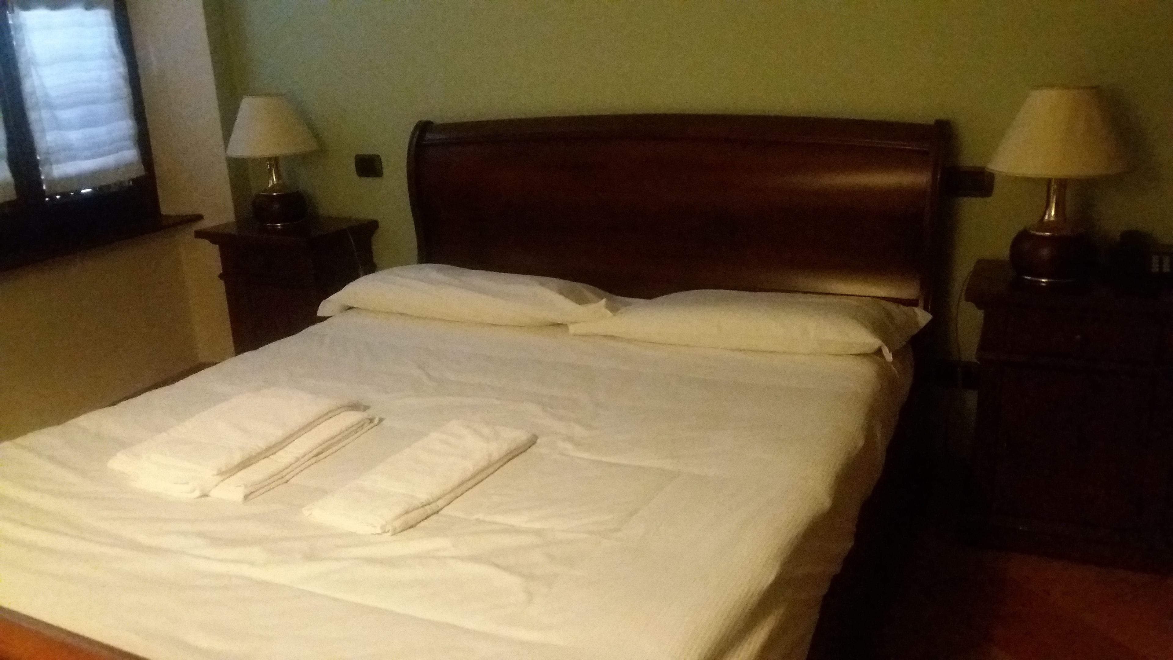 A neatly made bed with pillows and folded towels, two bedside lamps, and a window with blinds.