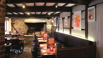 Interior acogedor de un restaurante con paredes de piedra, vigas de madera y una chimenea de piedra. Las mesas están preparadas con menús y velas.