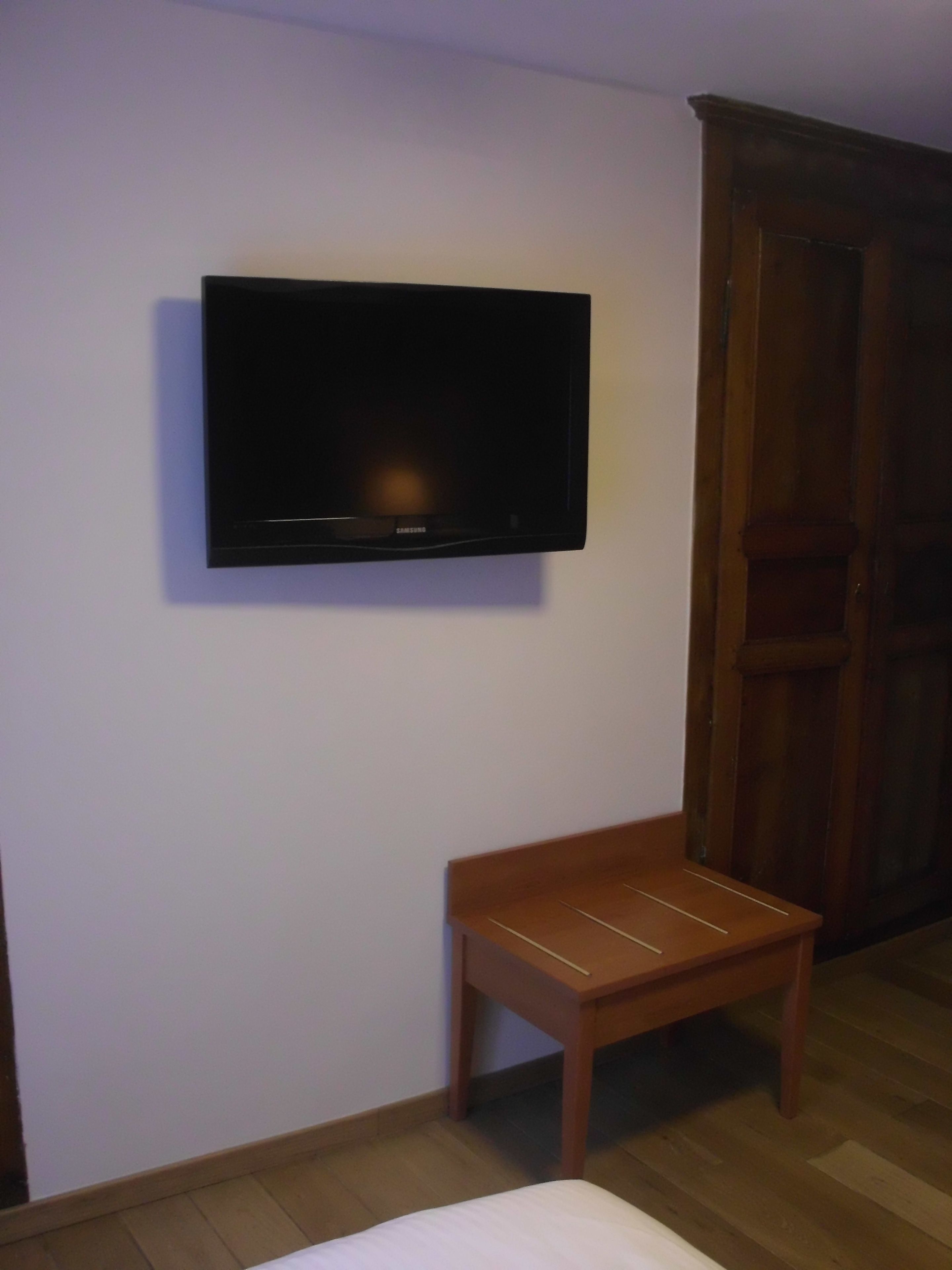 A room with a television mounted on the wall, a wooden bench, and a wooden wardrobe.