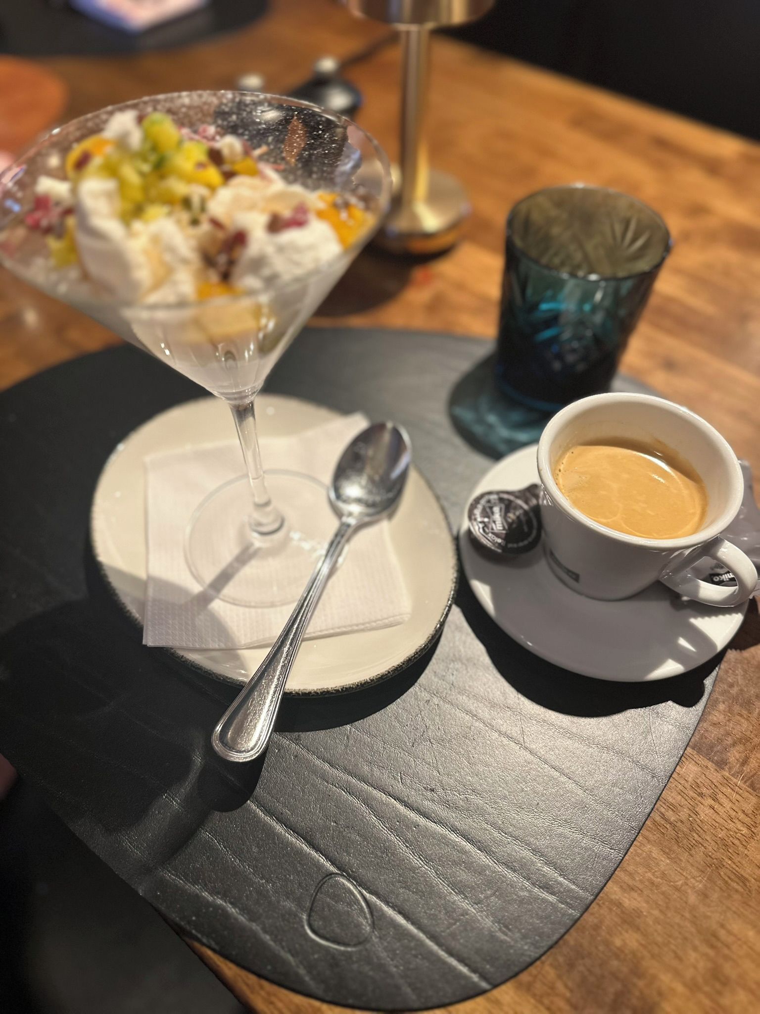 Ein Dessert in einer Glaskugel, ein Löffel, eine Tasse Kaffee und ein Glas Wasser auf einem Tisch.