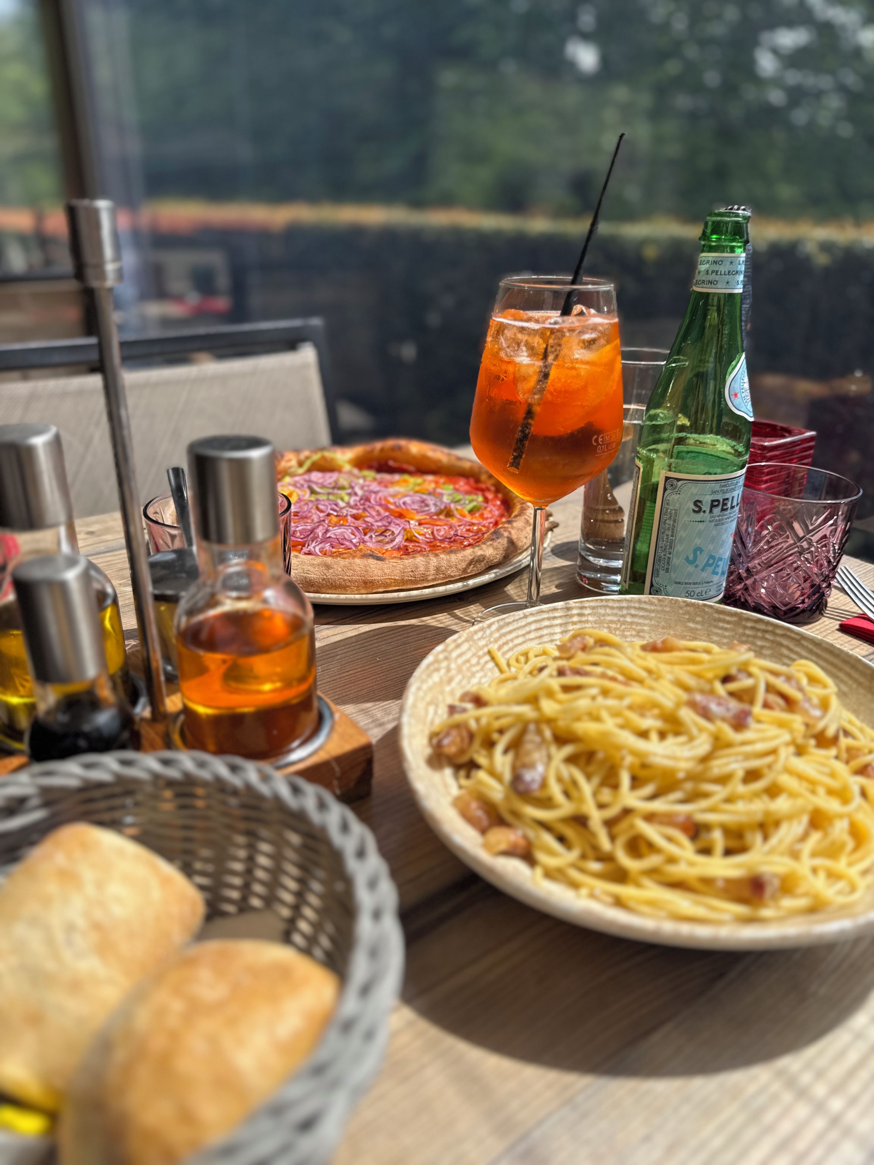 Un tavolo con una pizza, un bicchiere di bevanda, una bottiglia di olio d'oliva e un piatto di spaghetti.