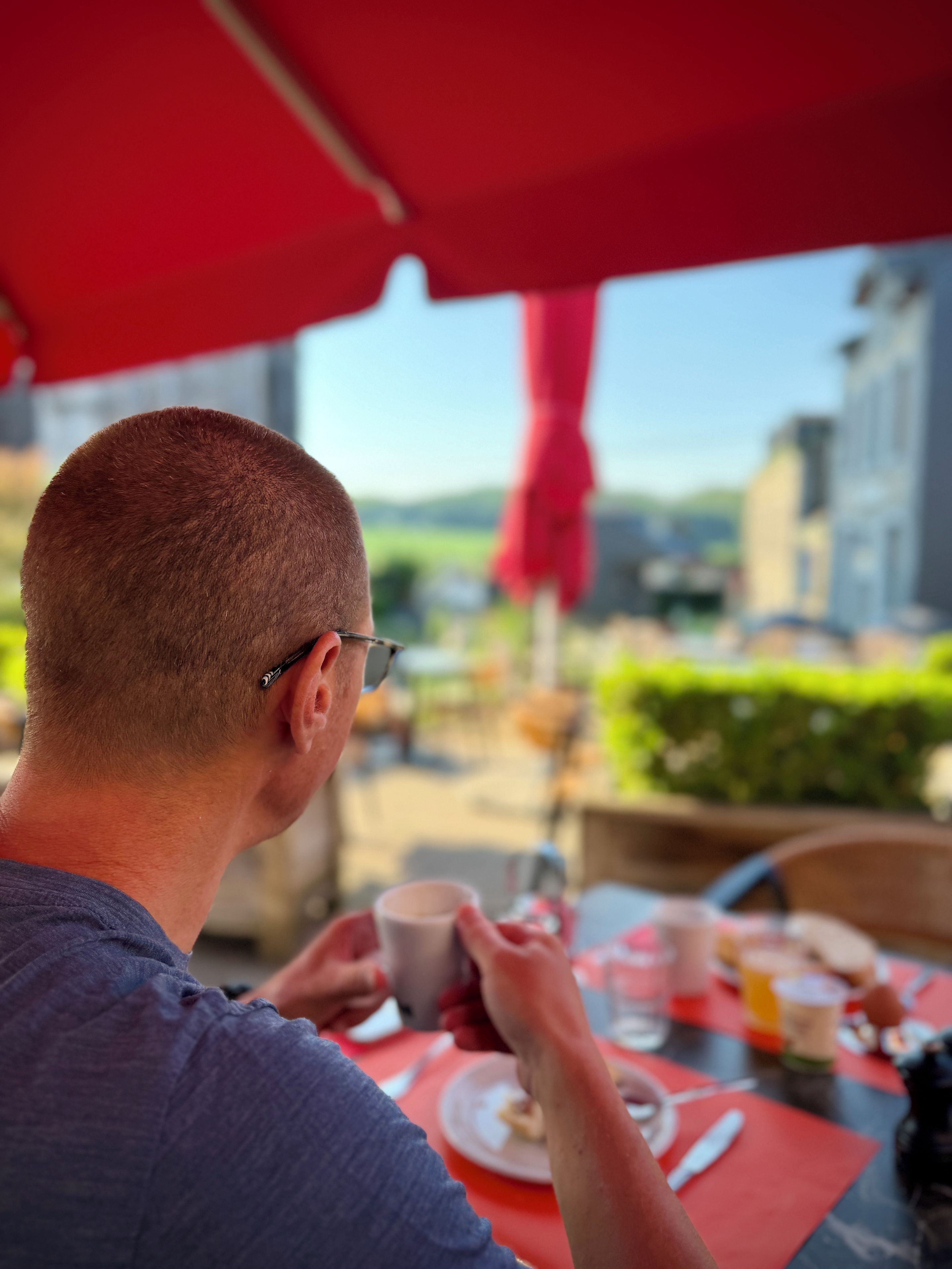 Une personne est assise à une table à l’extérieur, sous un parasol rouge, avec une tasse et une assiette remplie de nourriture.