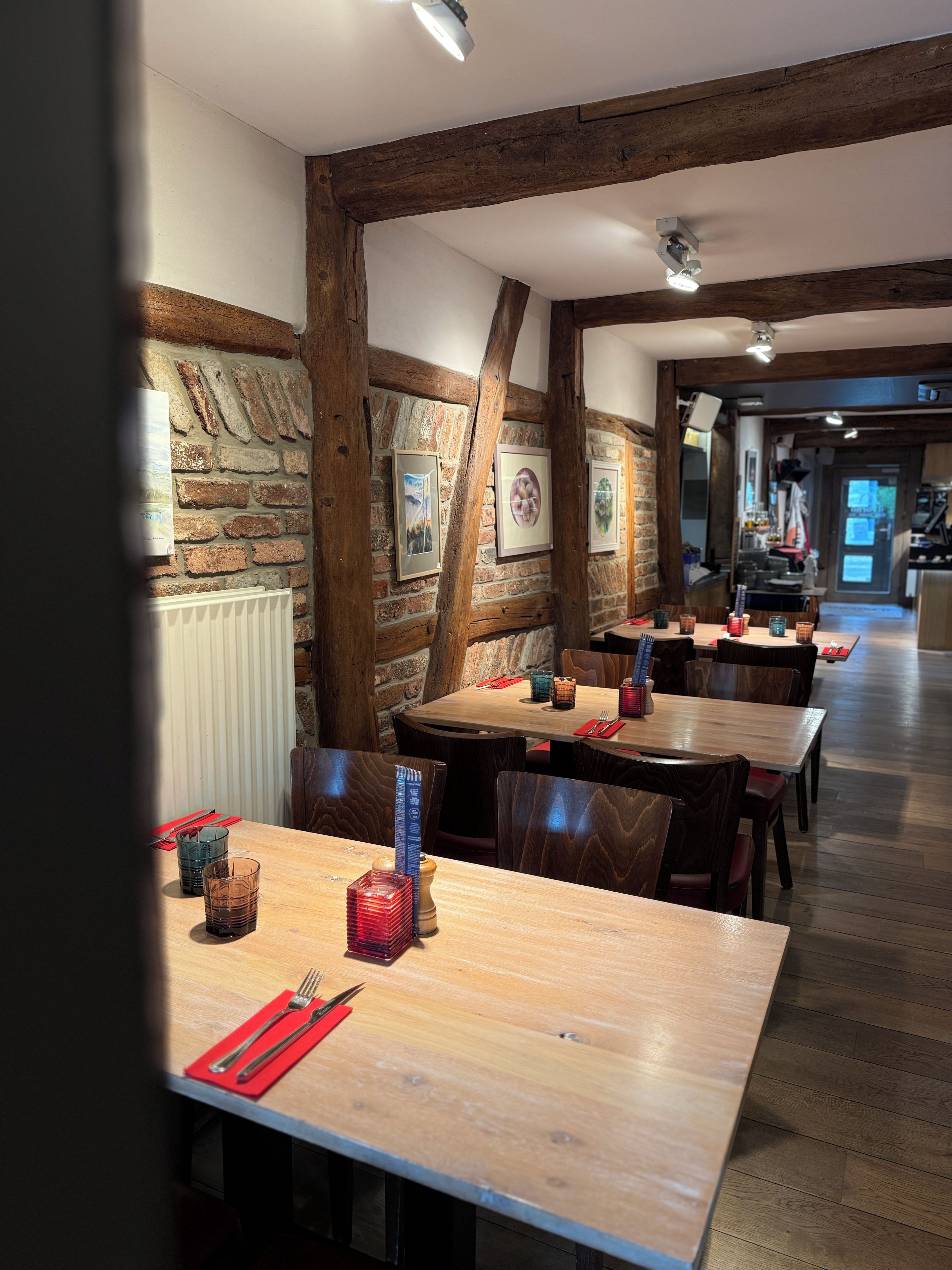 Interior de restaurante con vigas de madera, paredes de ladrillo y cuadros enmarcados. Mesas dispuestas con servilletas rojas y copas.