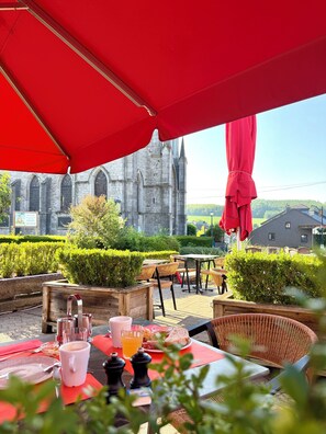 Un espace de restauration en plein air avec des tables et des chaises, un parasol rouge et, en arrière-plan, une vue sur une église historique.
