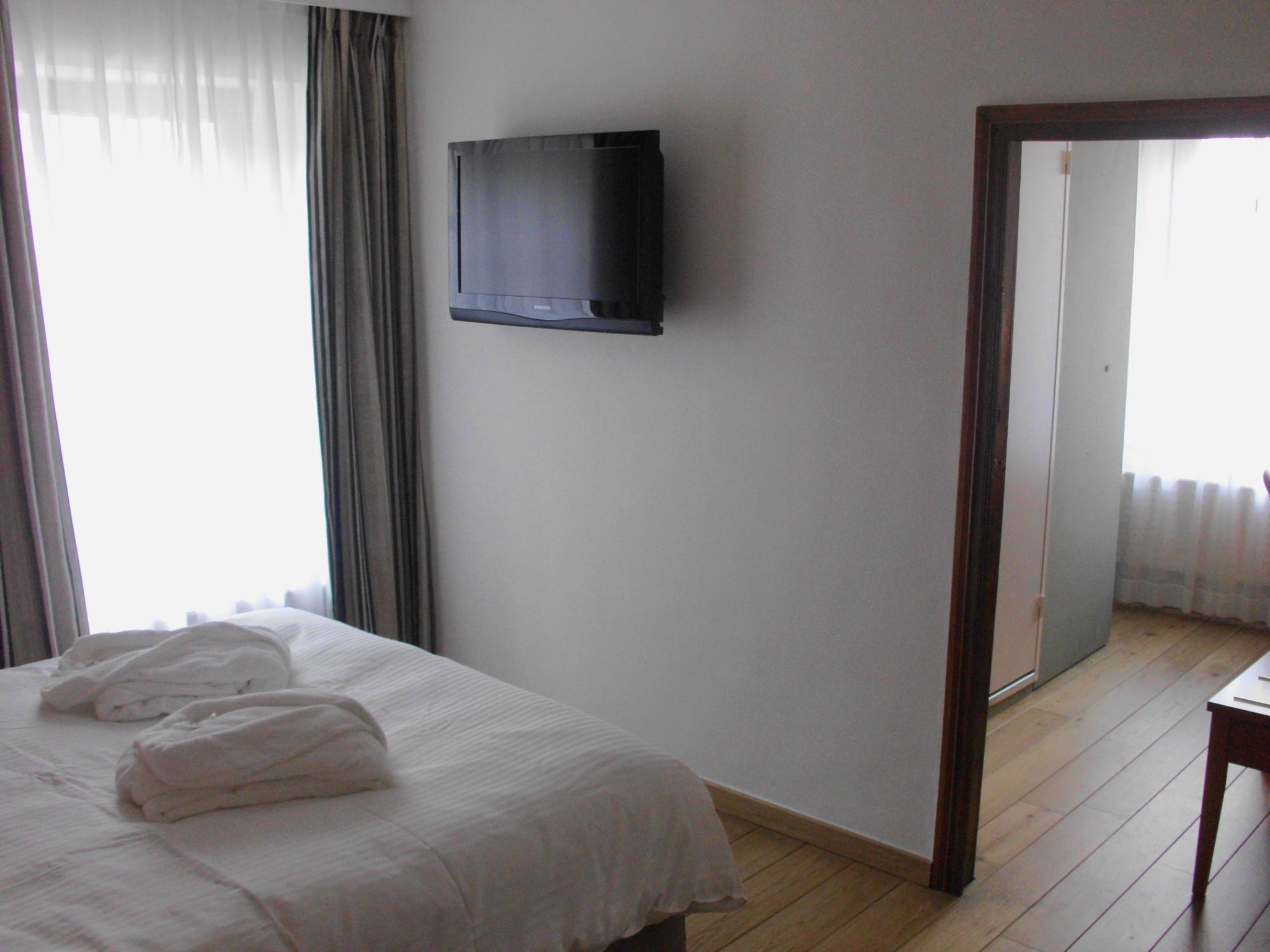 A hotel room with a bed, a television mounted on the wall, and a wooden floor.