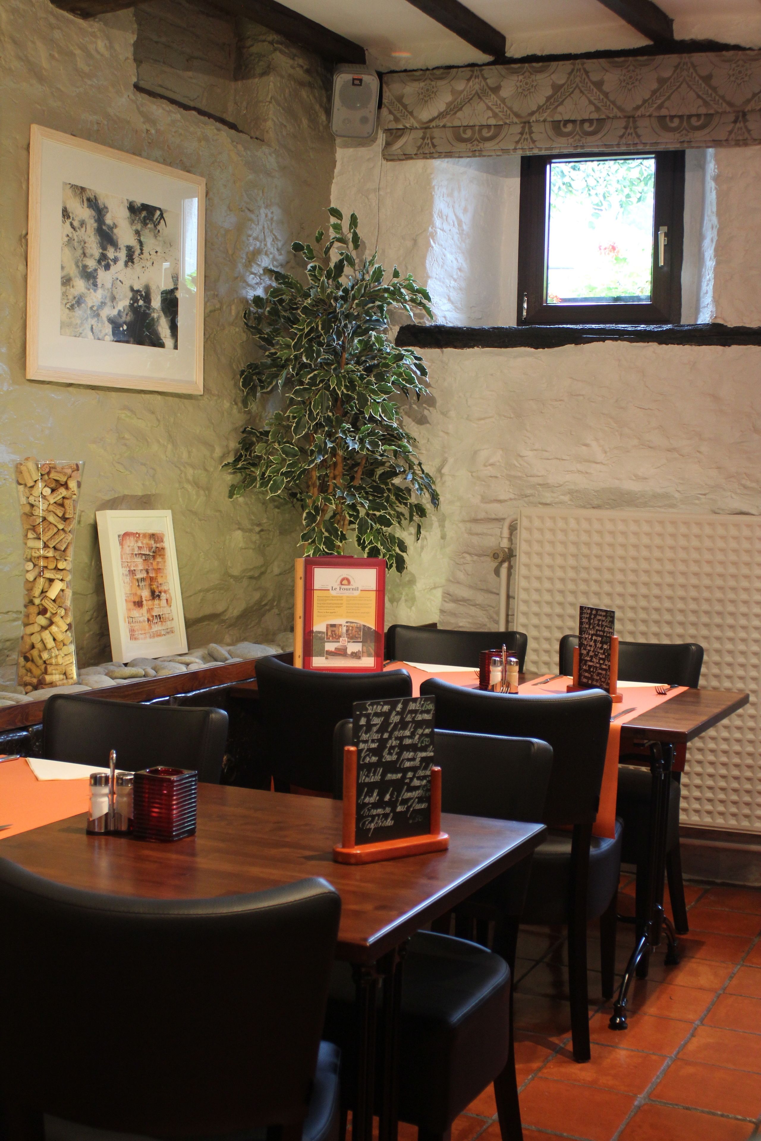 A restaurant interior with wooden tables, black chairs, a large potted plant, framed artwork, and a chalkboard menu.