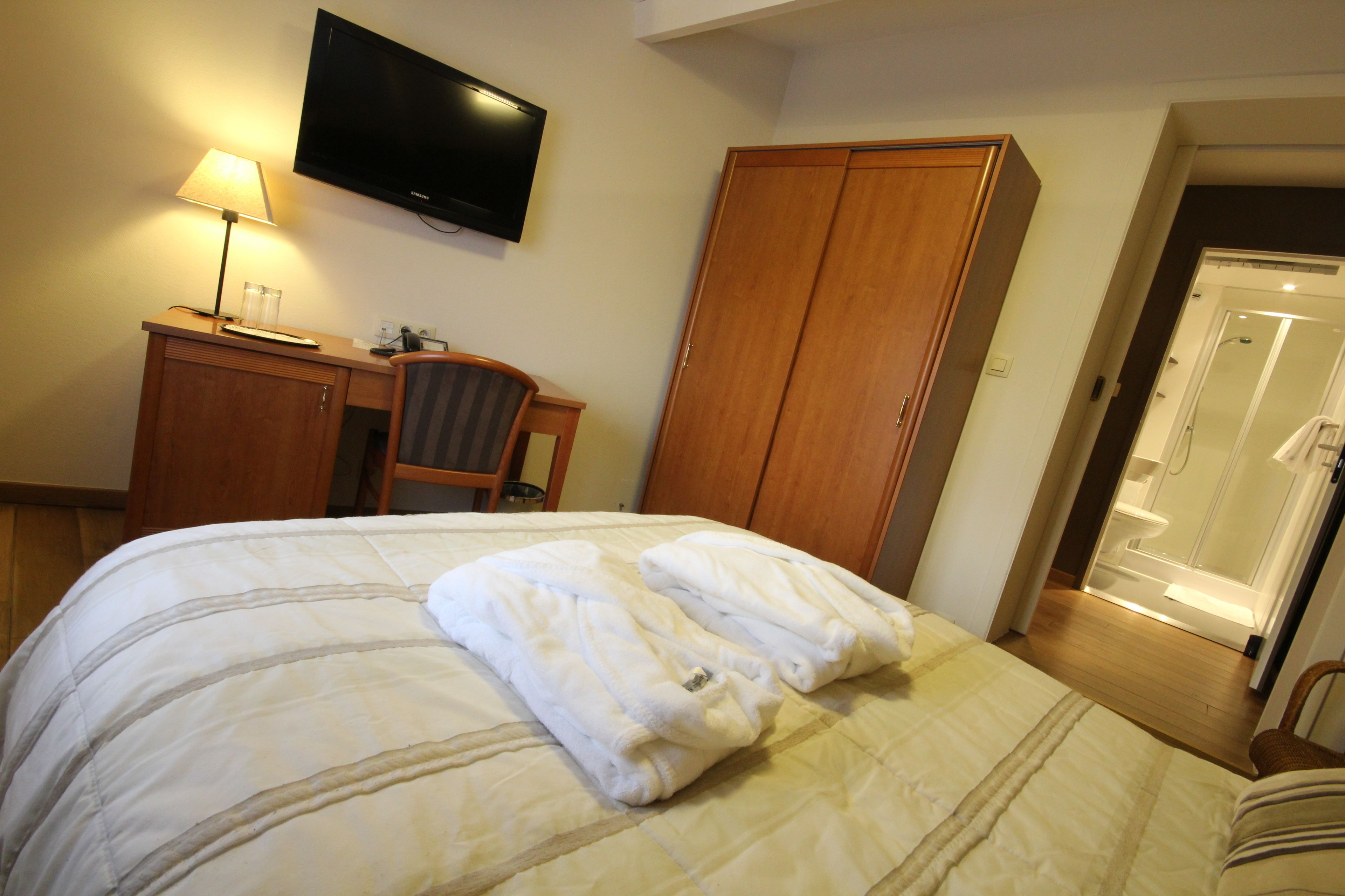A hotel room with a bed, a desk with a lamp, a TV mounted on the wall, and a wardrobe.