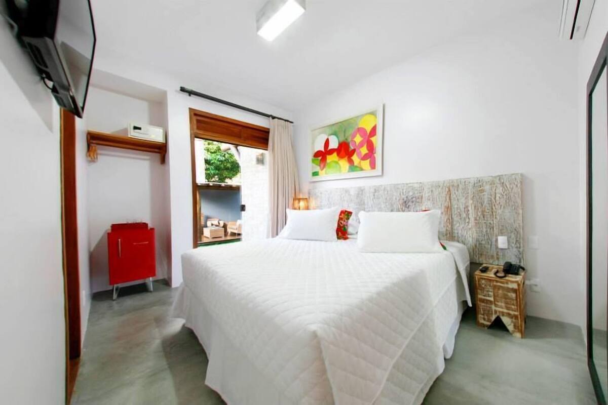 A hotel room with a large bed, a red cabinet, a small wooden side table, and a painting on the wall.