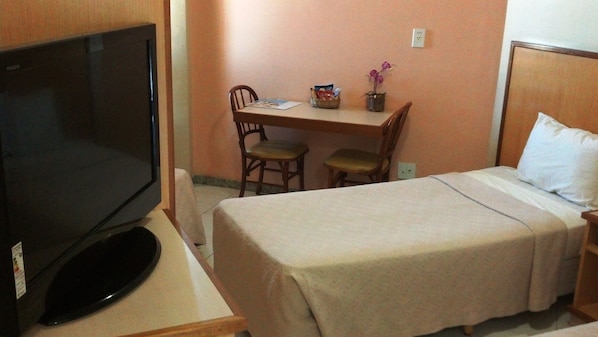 Habitación de hotel con cama, televisor, escritorio con dos sillas, mesita con una cesta y una planta en maceta.