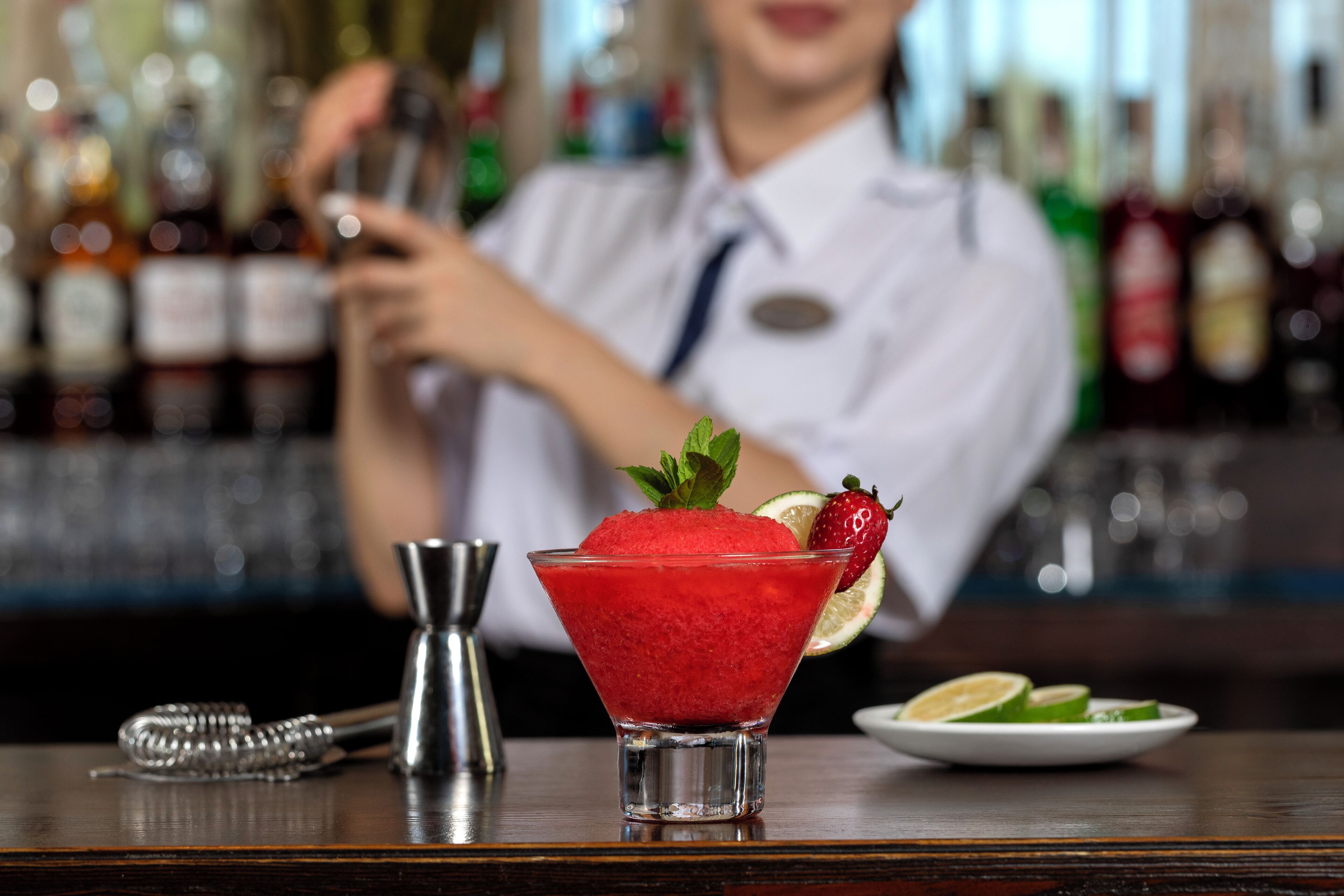 Un còctel en un vas de martini amb una fresa i una llima com a guarnició, amb un agitador de còctels i un plat amb llesques de llima sobre la barra.