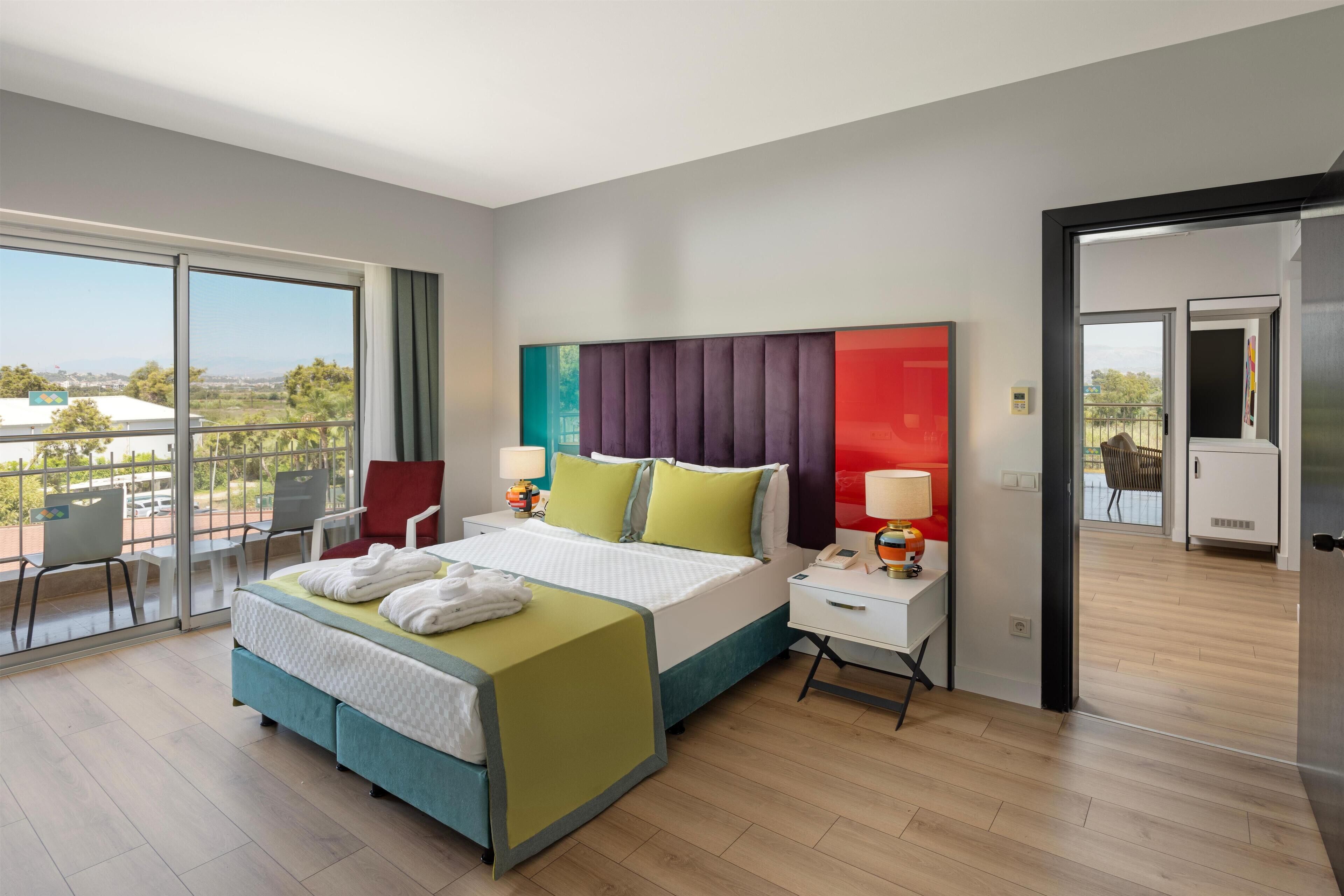 A modern hotel room with a large bed, a desk, and a balcony with chairs.