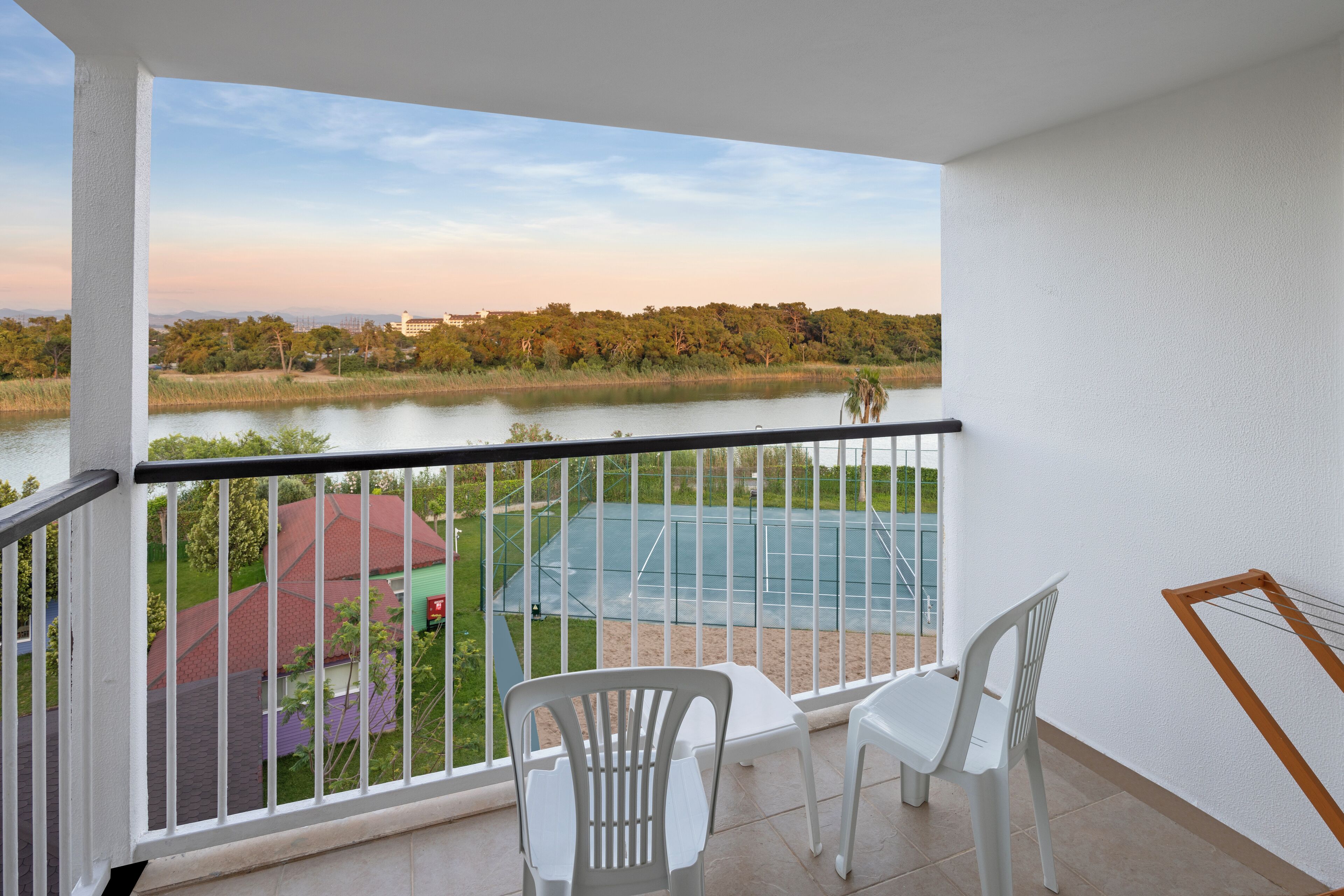A balcony with a view of a lake, a tennis court, and a green building in the background.