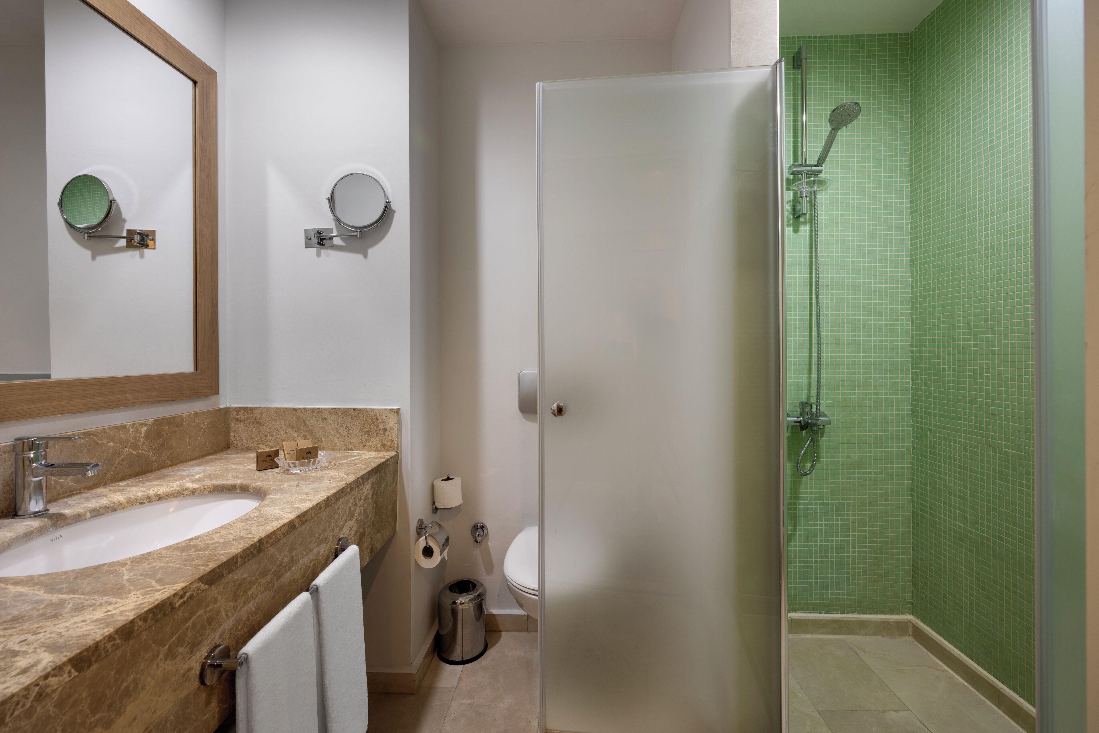 Un bagno con un lavabo in marmo, due specchi rotondi, un water e una doccia con piastrelle verdi.