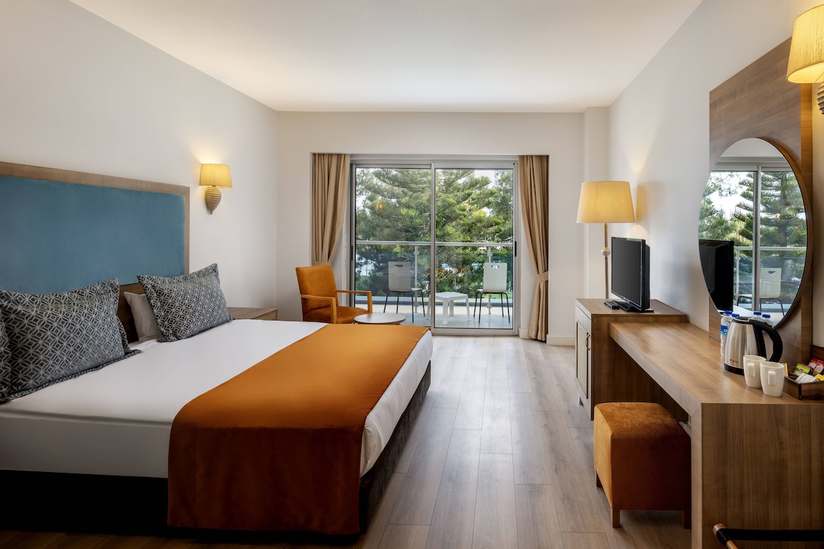 A hotel room with a bed, a desk with a TV, a chair, a small table, and a balcony with a view of trees.