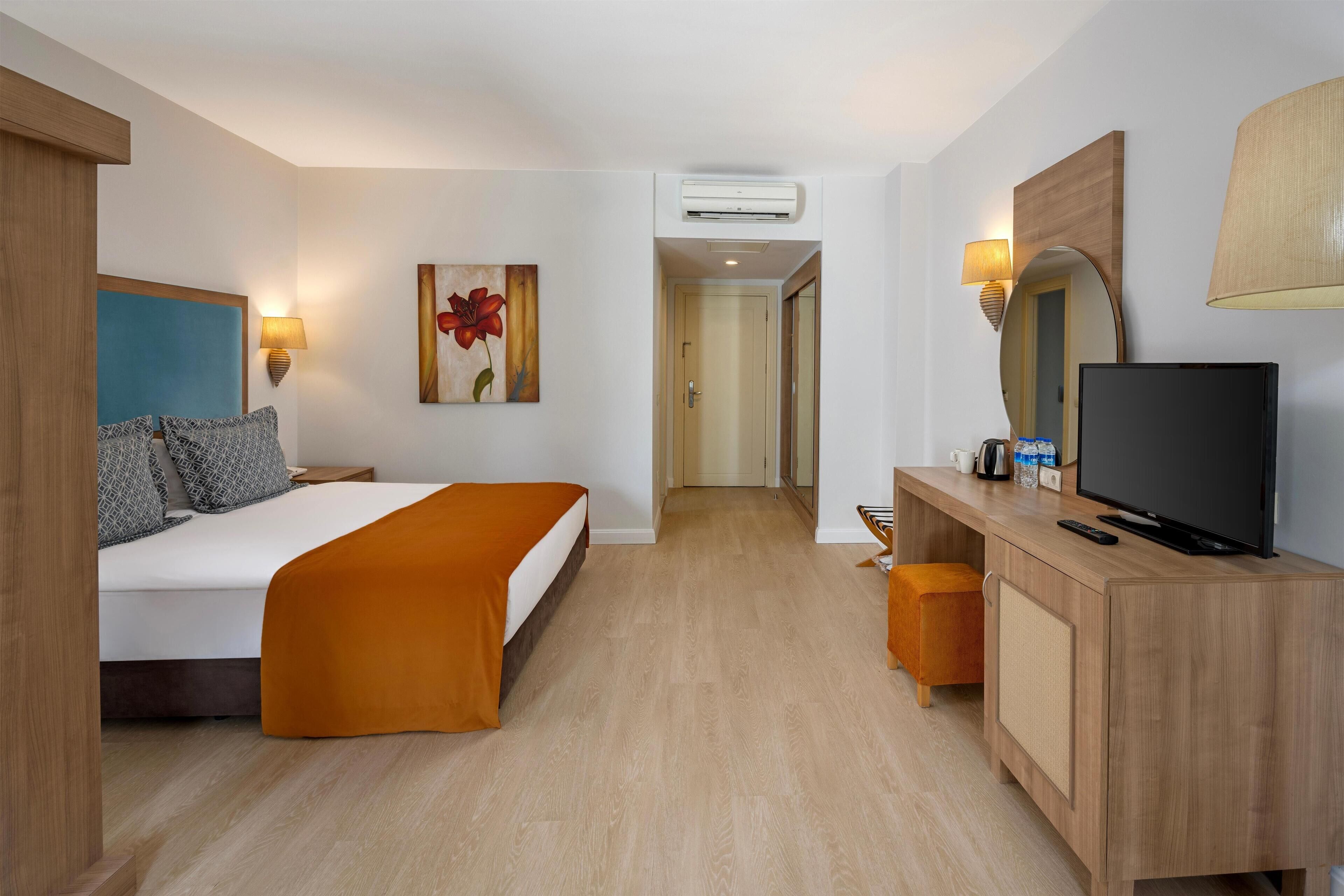 A hotel room with a bed, a desk with a television, a mirror, and a painting on the wall.