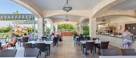 Un espacio de comedor al aire libre con mesas y sillas, una barra con una persona atendiendo, y una vista de un parque infantil al fondo.