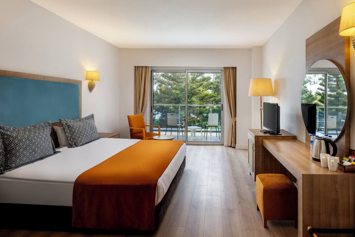 A hotel room with a large bed, a desk with a television, and a balcony with a view of trees.