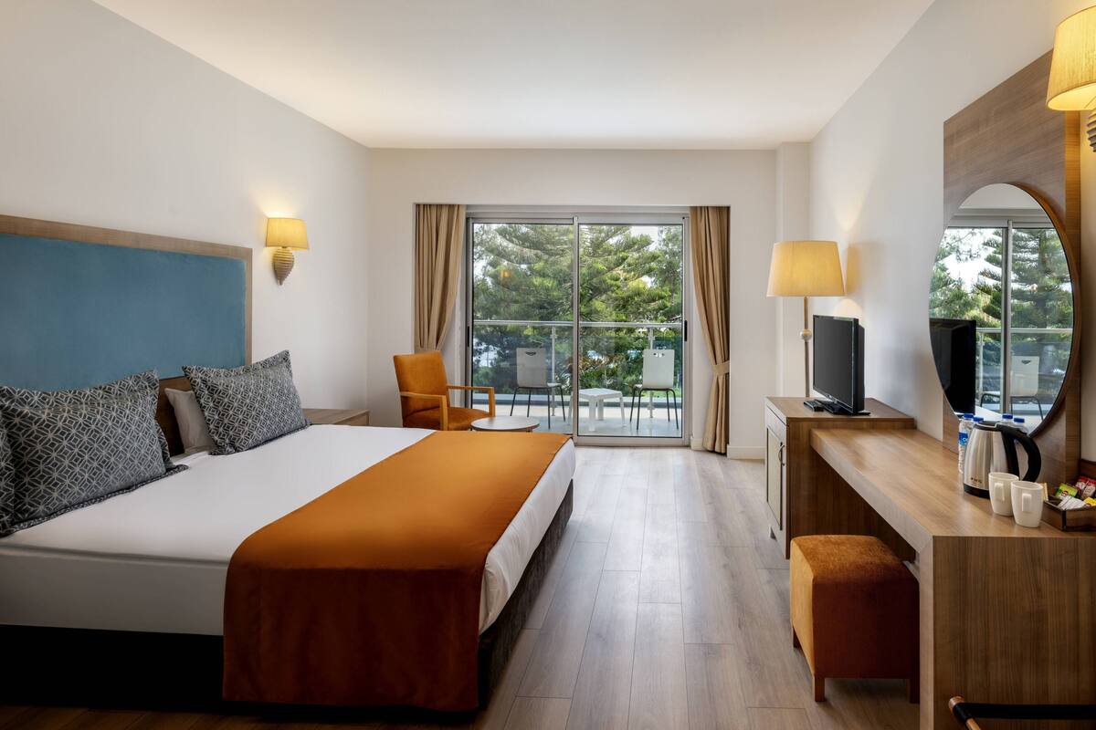 Quarto de hotel com cama, escrivaninha com televisão, cadeira, mesa pequena e varanda com vista para árvores.
