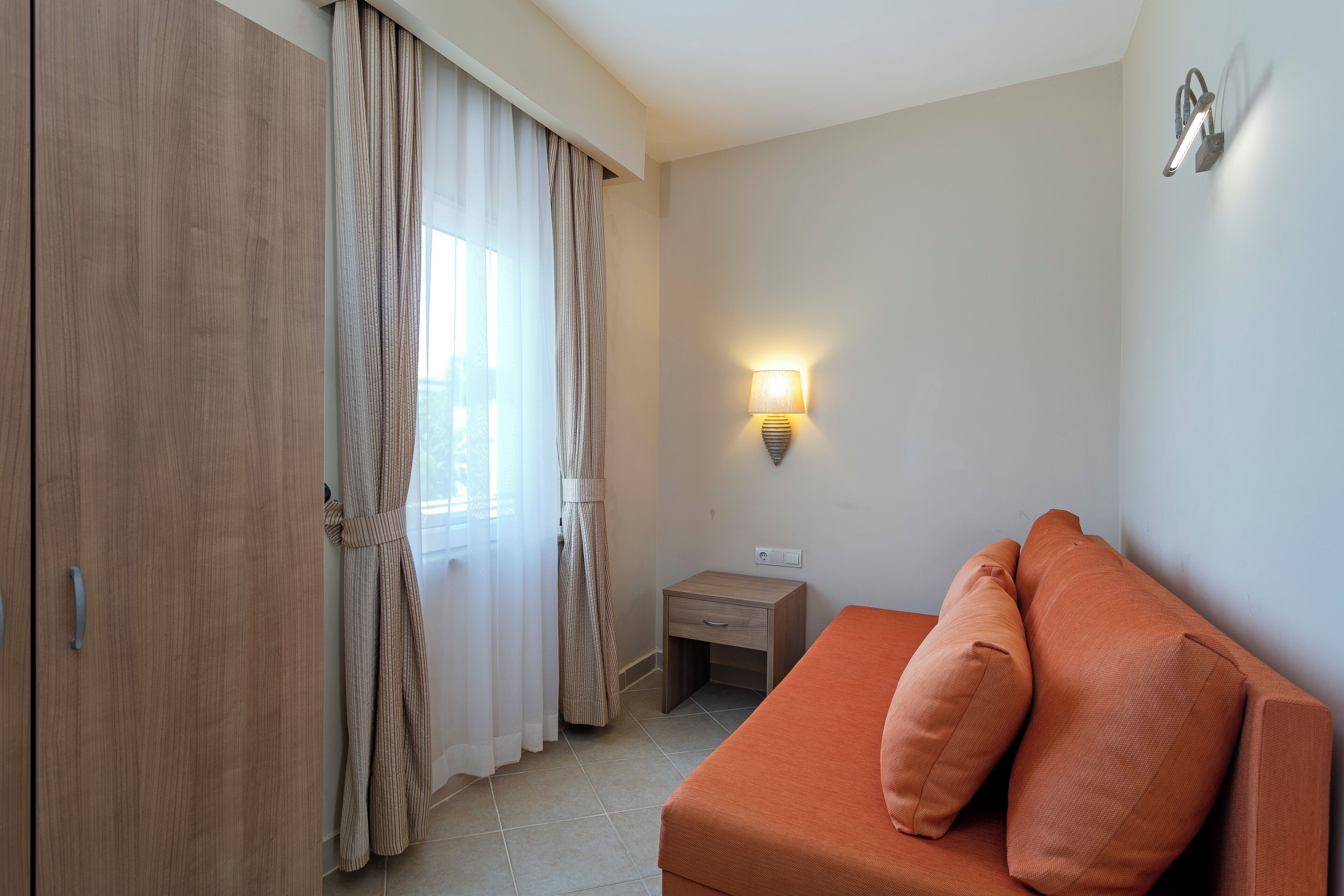 A room with a wooden wardrobe, a bed with two pillows, a nightstand, a wall-mounted lamp, and a window with curtains.