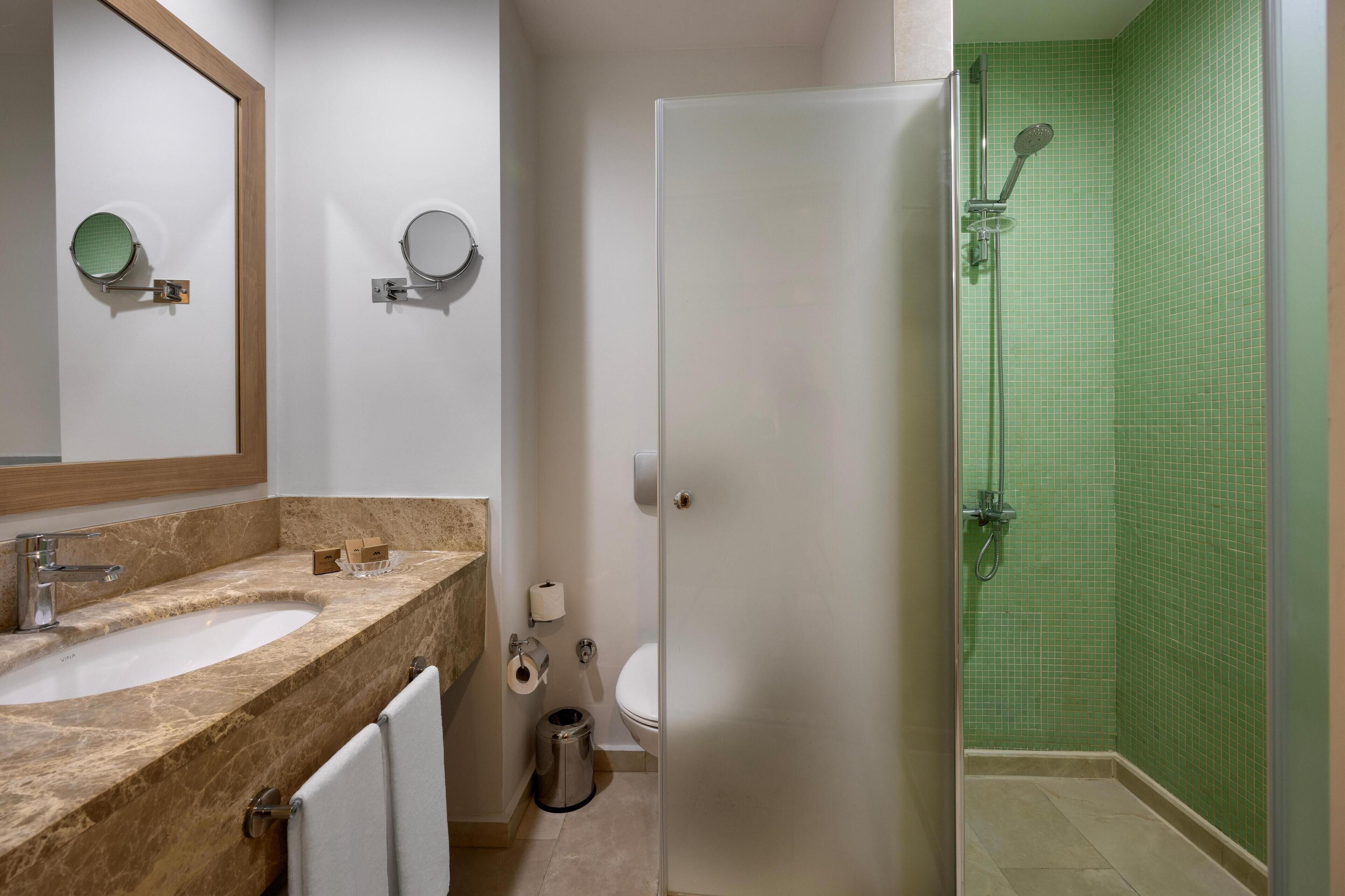 A bathroom with a marble sink, two mirrors, a toilet, and a green-tiled shower area.