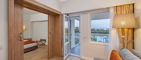 A hotel room with a wooden door, a bed, a balcony with a view, and a sofa with cushions.