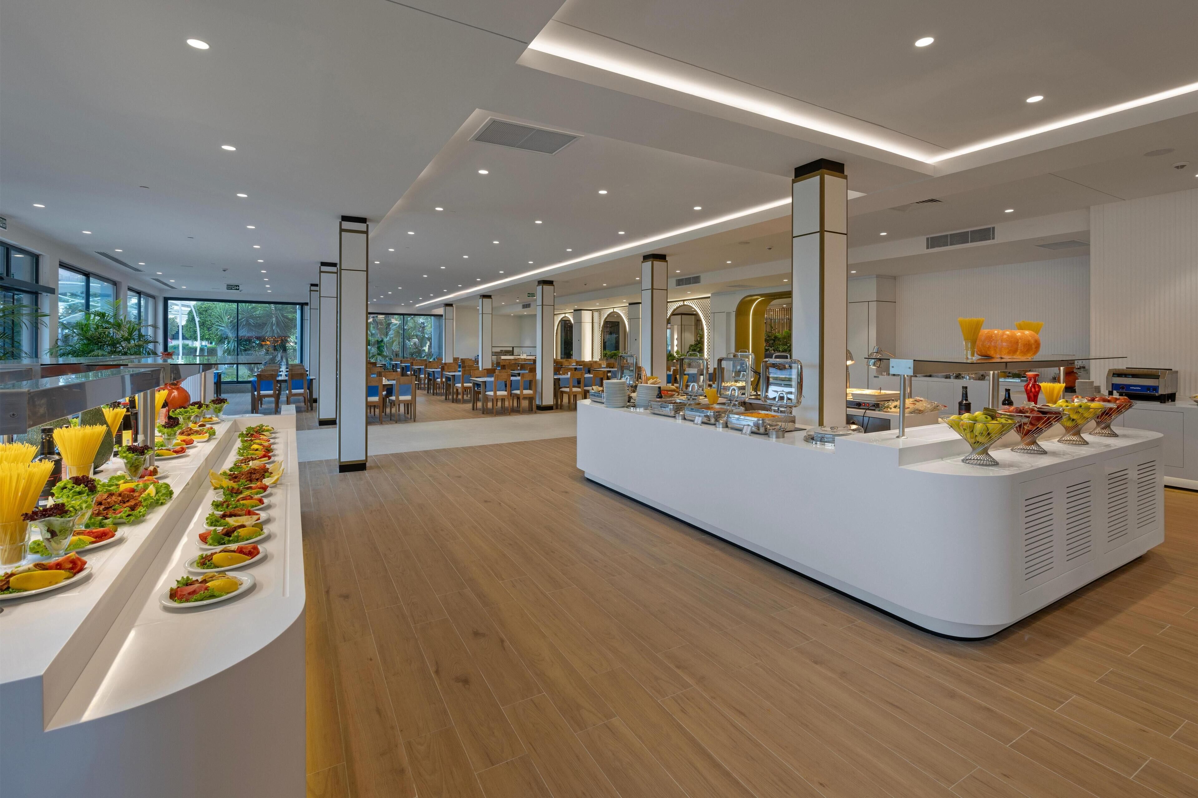 A modern hotel dining area with a long buffet table, multiple seating areas, and large windows providing natural light.