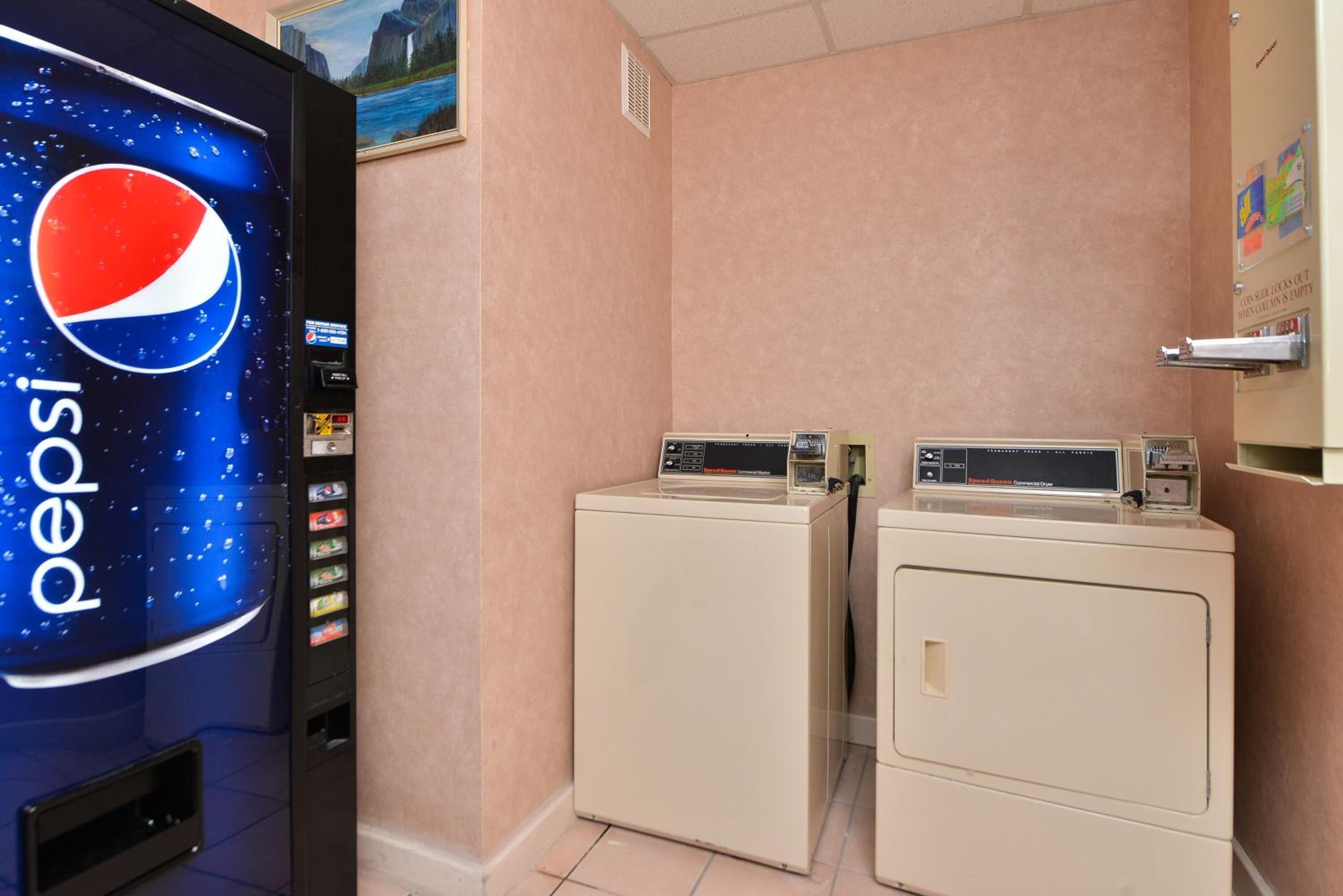 Una habitación con una máquina expendedora de Pepsi, dos lavadoras y un tablón de anuncios en la pared.