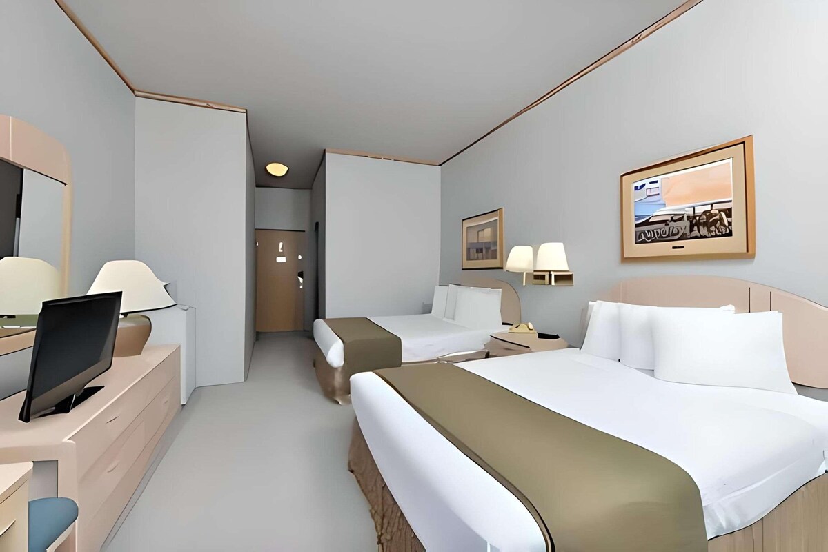 Una habitación de hotel con dos camas, un escritorio con computadora, un televisor y un cuadro en la pared.