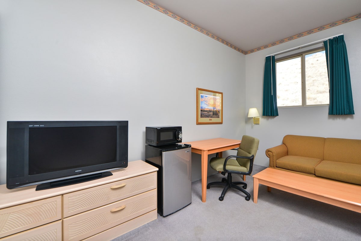 A room with a flat-screen TV, a microwave, a refrigerator, a desk, a chair, a sofa, a framed picture on the wall, and a window with curtains.